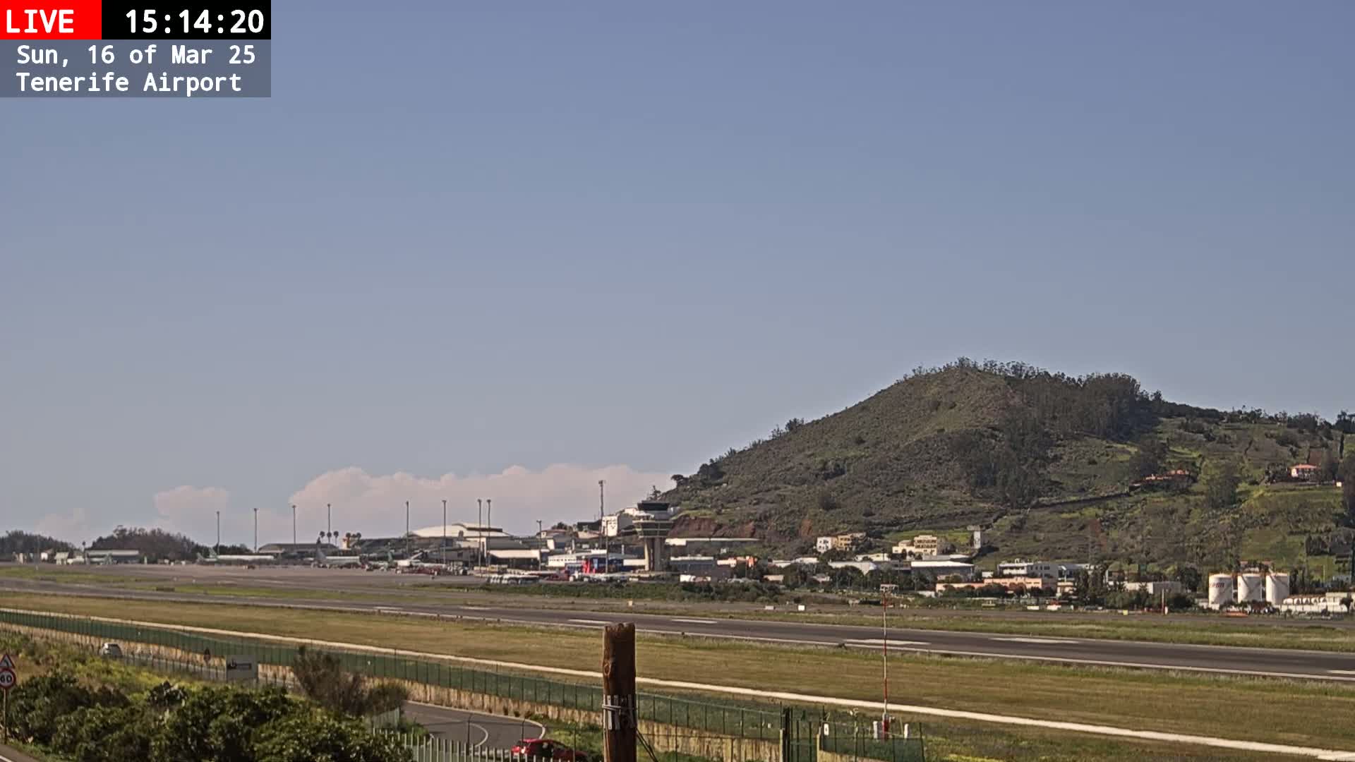 A sunny day at an airport shows a runway, terminal buildings, and a hill in the background with some scattered clouds.