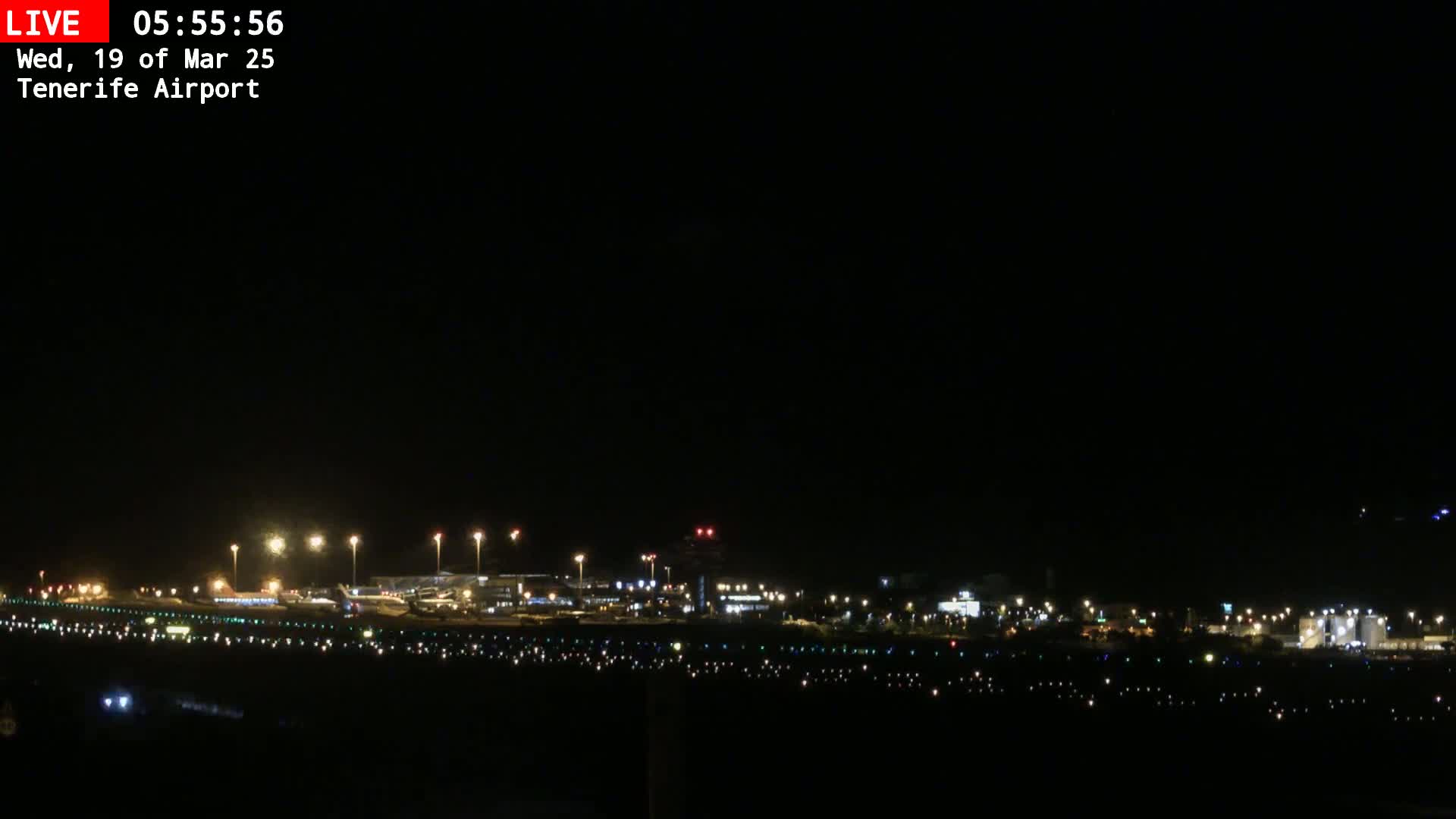 A nighttime long shot of an airport runway and terminal buildings illuminated by lights under a clear, dark sky.