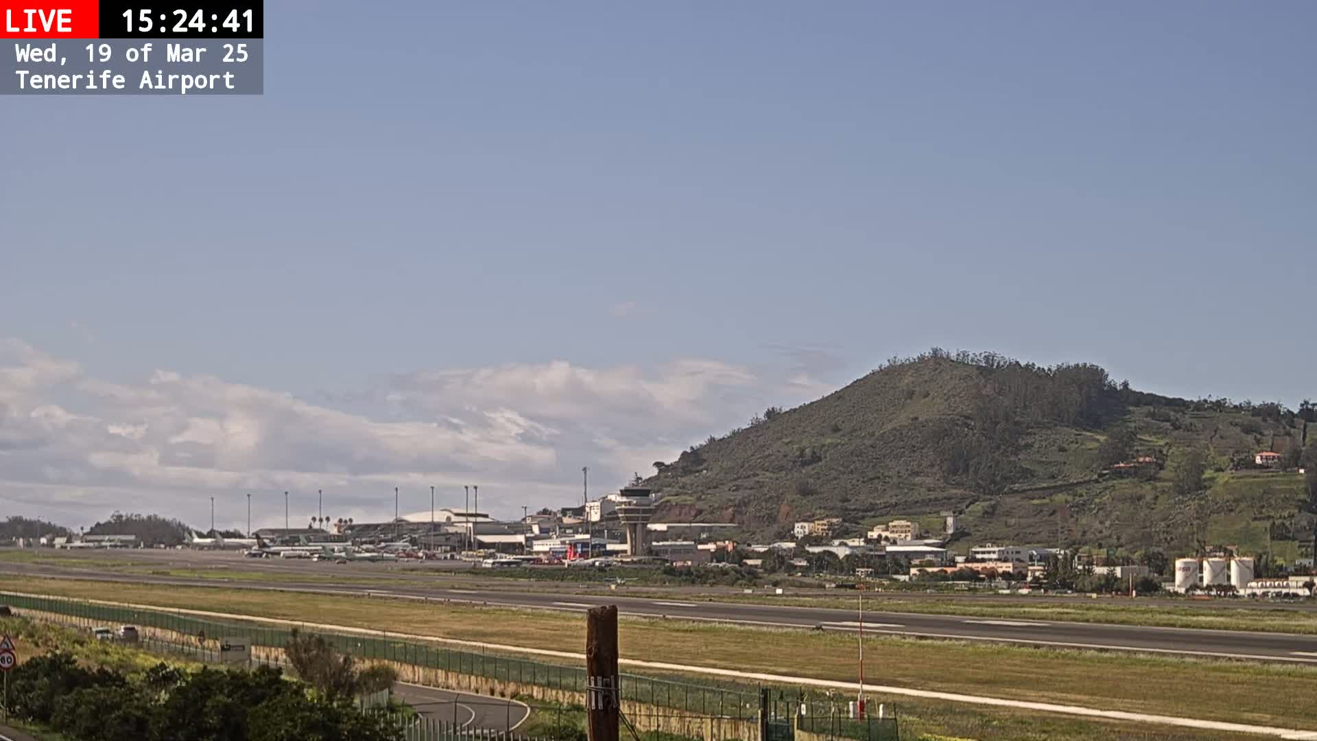 A mostly sunny day reveals a view of an airport runway, terminal buildings, and a large, green hill in the background.