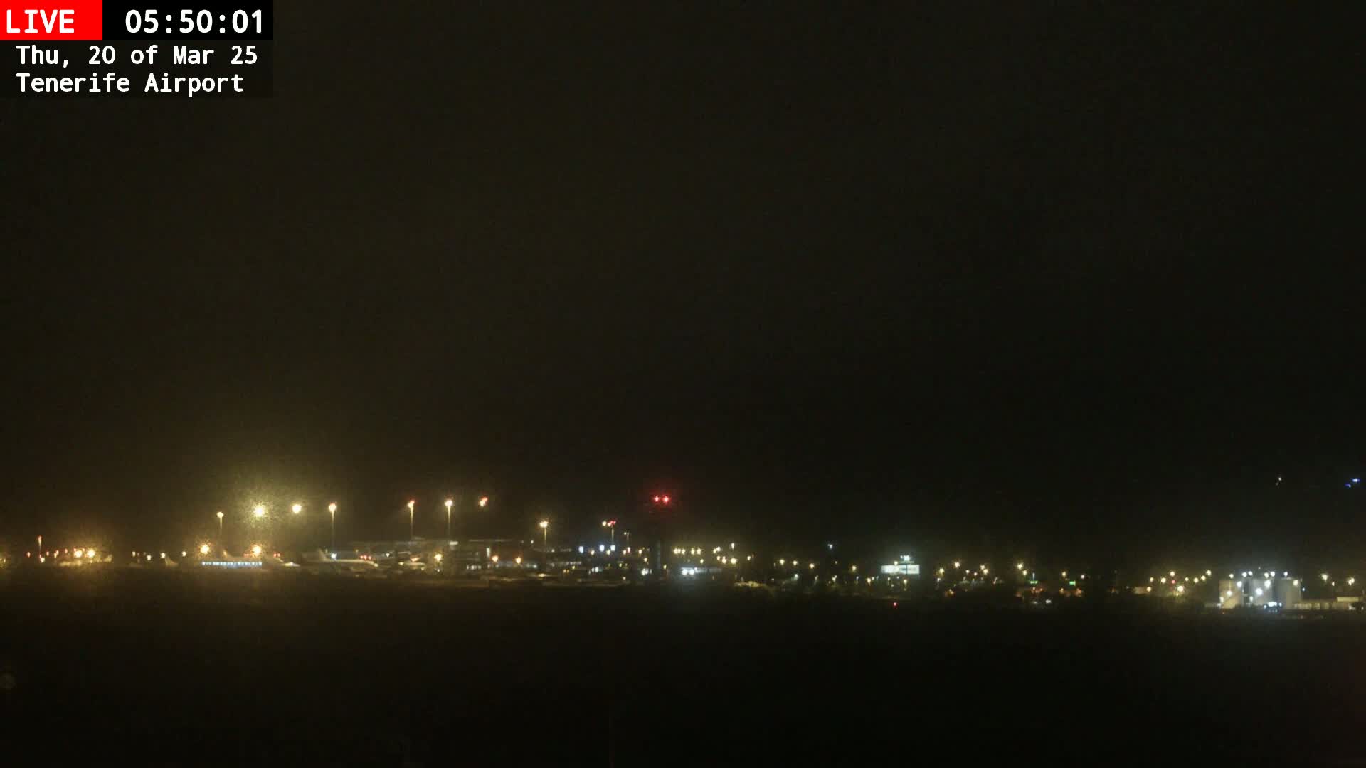 A nighttime view of an airport runway and terminal buildings, dimly lit under a dark, possibly overcast sky.