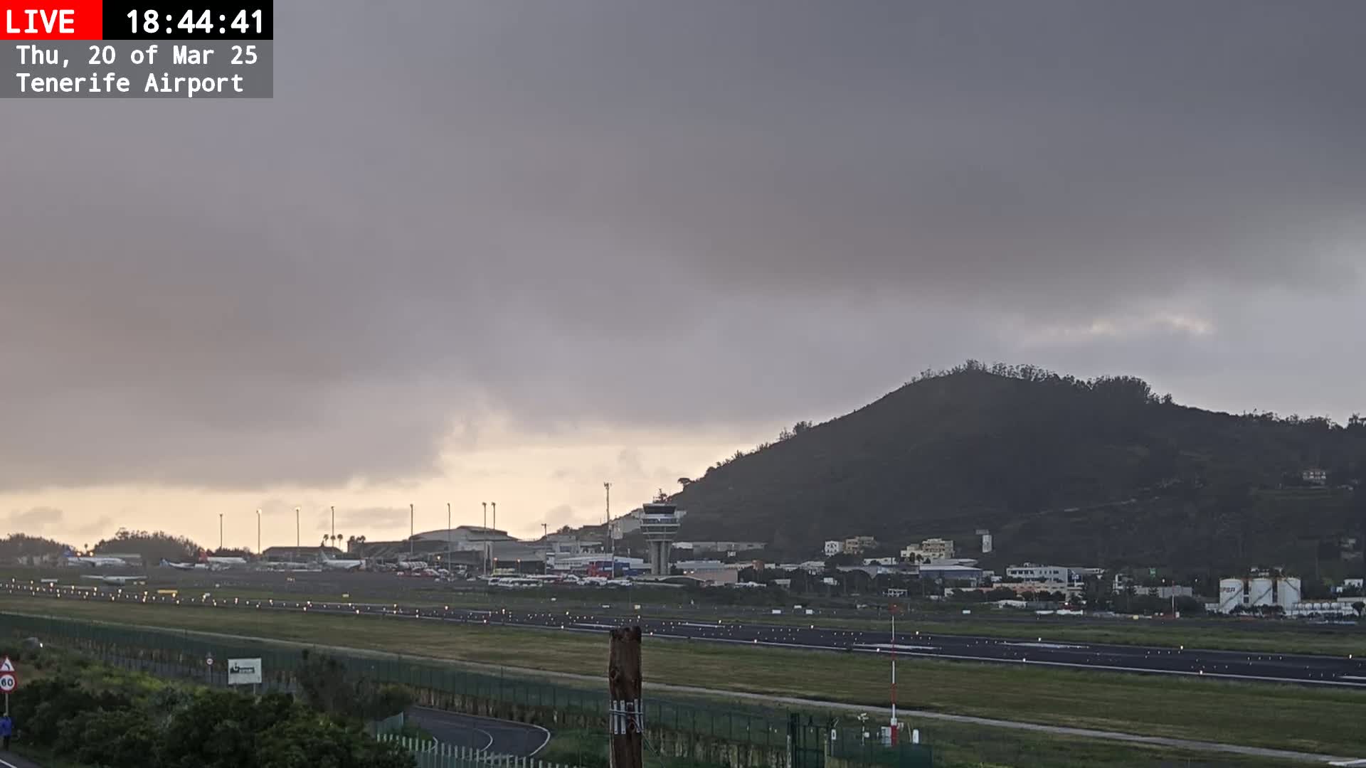 A view of an airport runway and terminal buildings under a cloudy, overcast sky, with a large hill in the background.