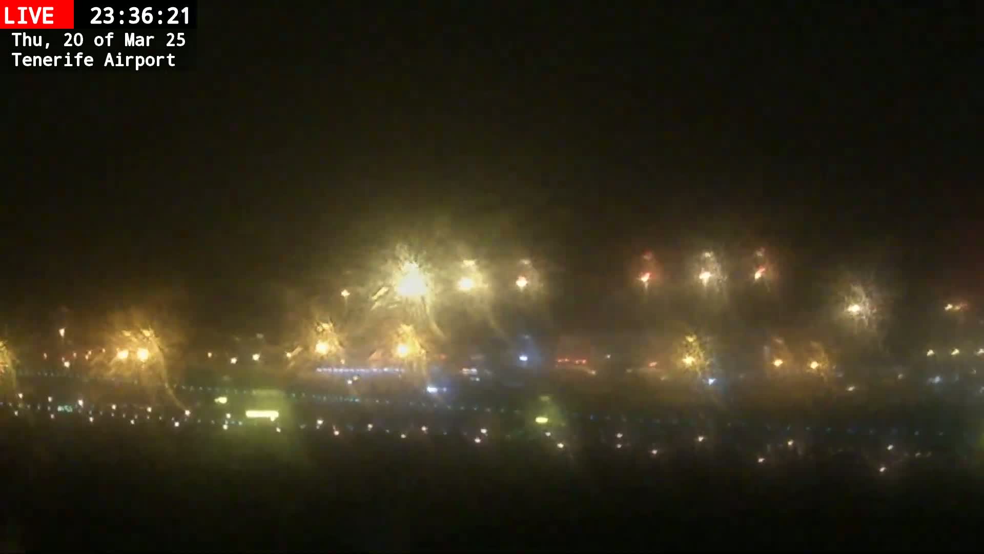 A nighttime, hazy view shows numerous lights along a long, low structure, possibly a road or runway, with some lights appearing brighter and more diffused than others.