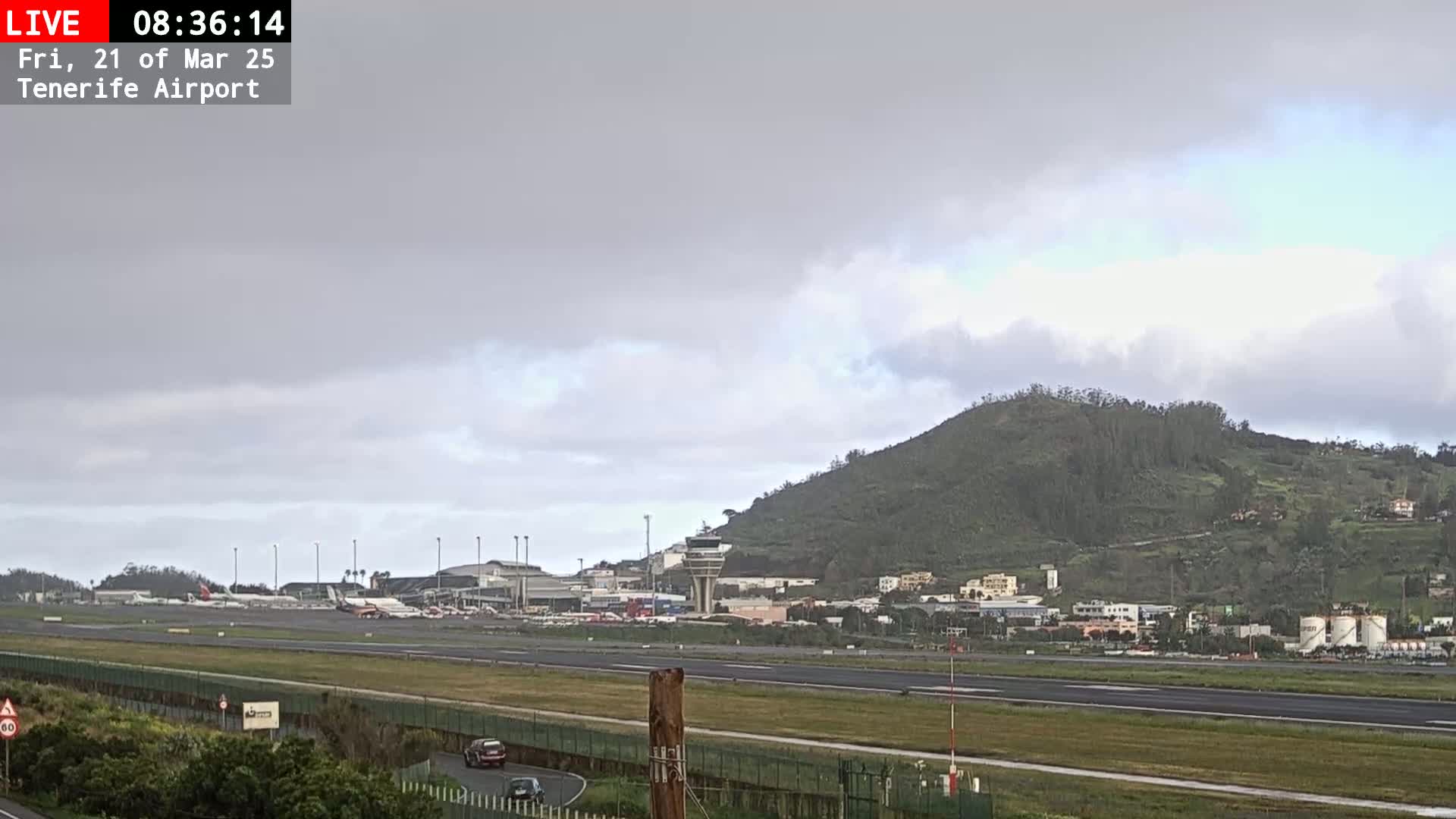 A view of an airport runway with several airplanes parked, a control tower, and a green hill in the background under an overcast sky.