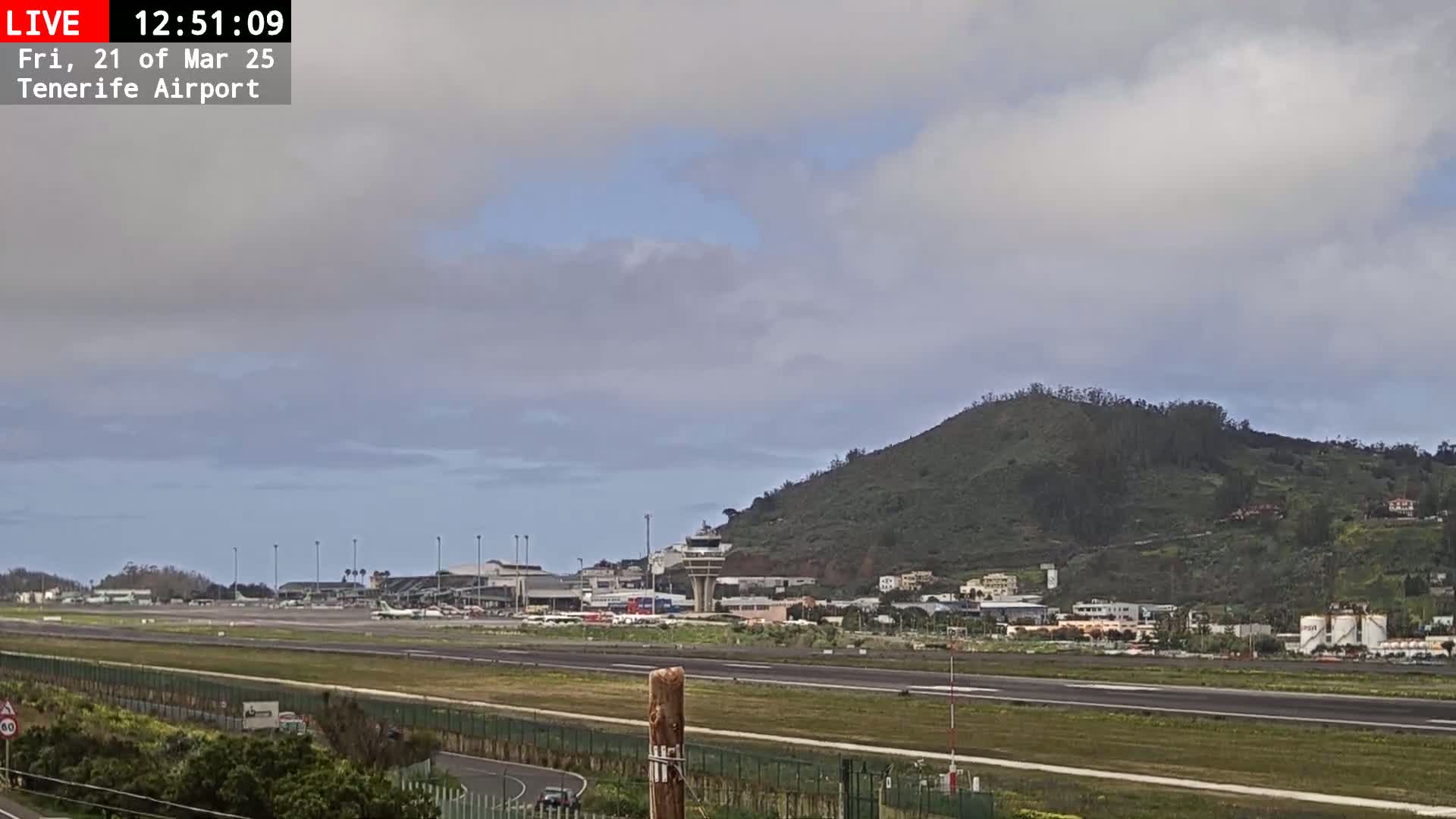 A mostly cloudy sky overlooks an airport runway with several planes visible, a control tower, and a green hill in the background.