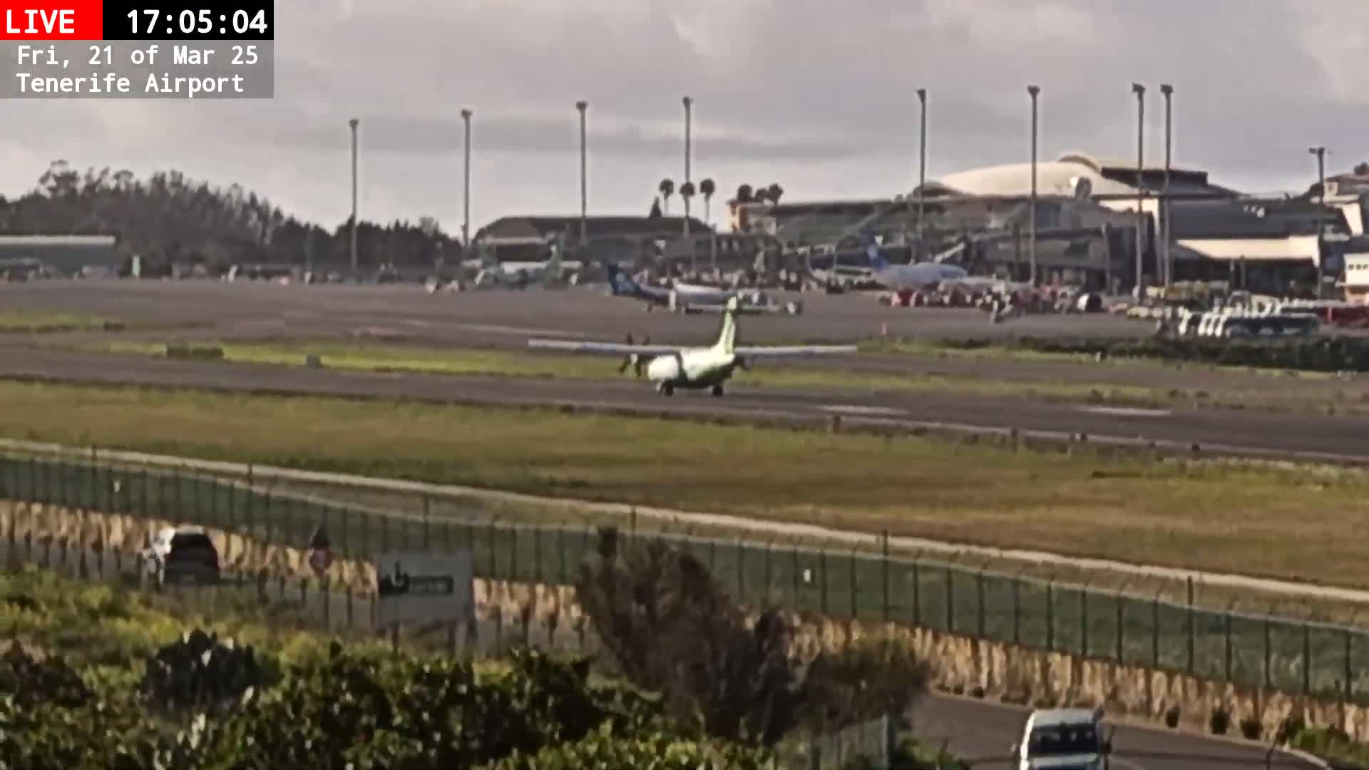 A small passenger plane taxis on an airport runway under overcast skies, with other planes and airport buildings visible in the background.