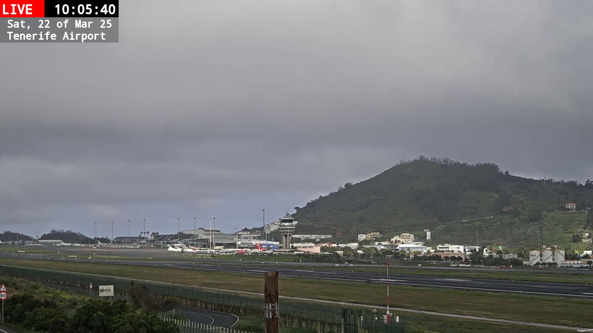 A view of an airport runway and terminal buildings under an overcast sky, with a large green hill in the background.