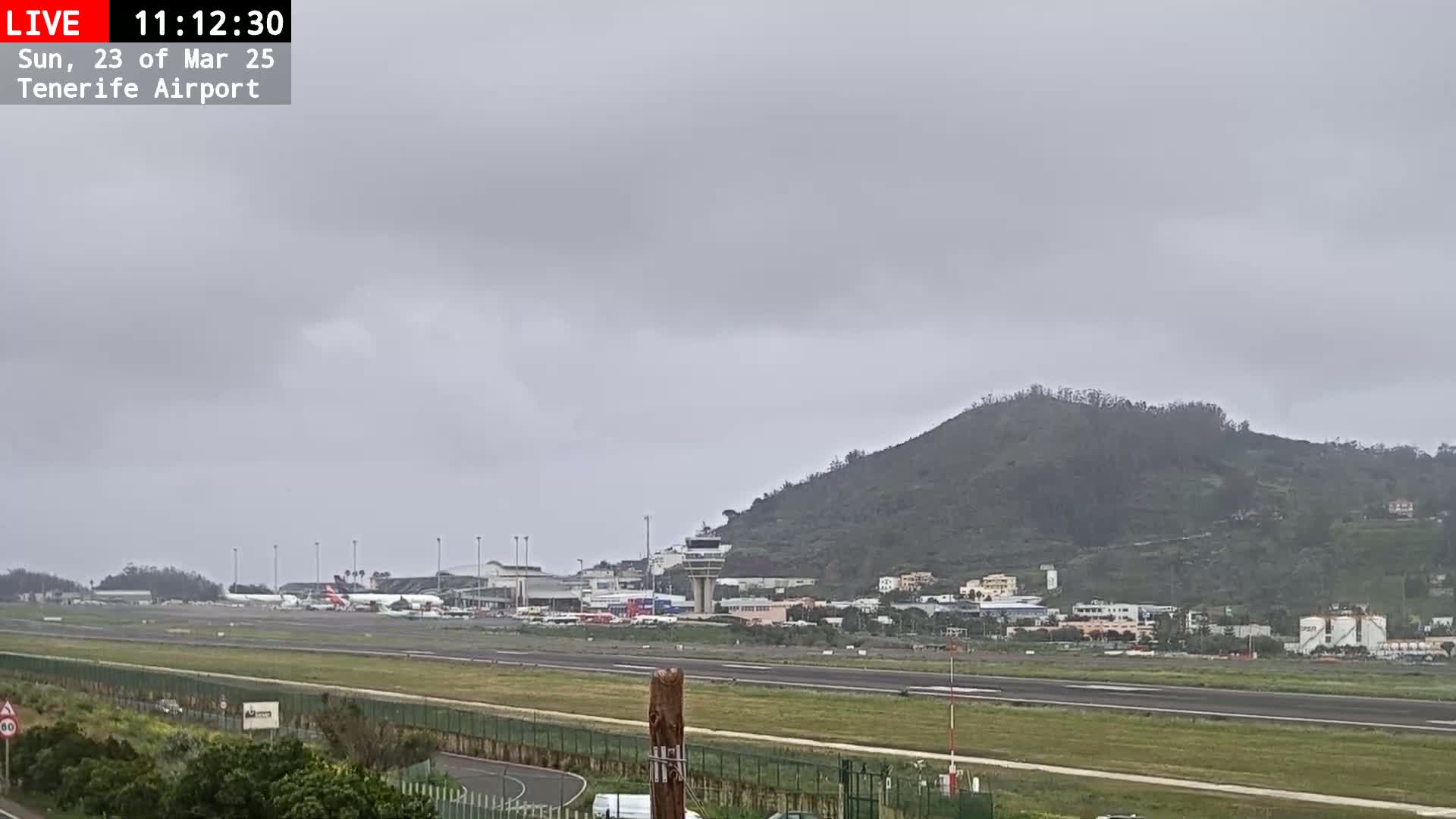 A view of an airport runway, terminal buildings, and a nearby hill under an overcast sky.