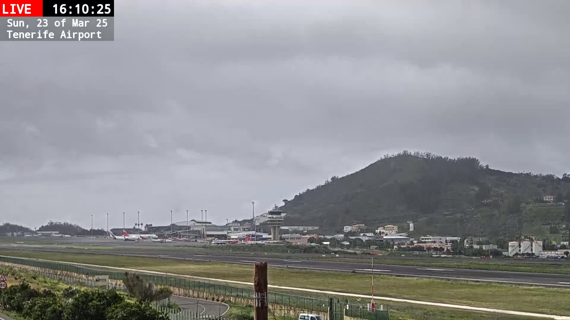 An airport runway and terminal are visible beneath an overcast sky, with a large hill in the background.