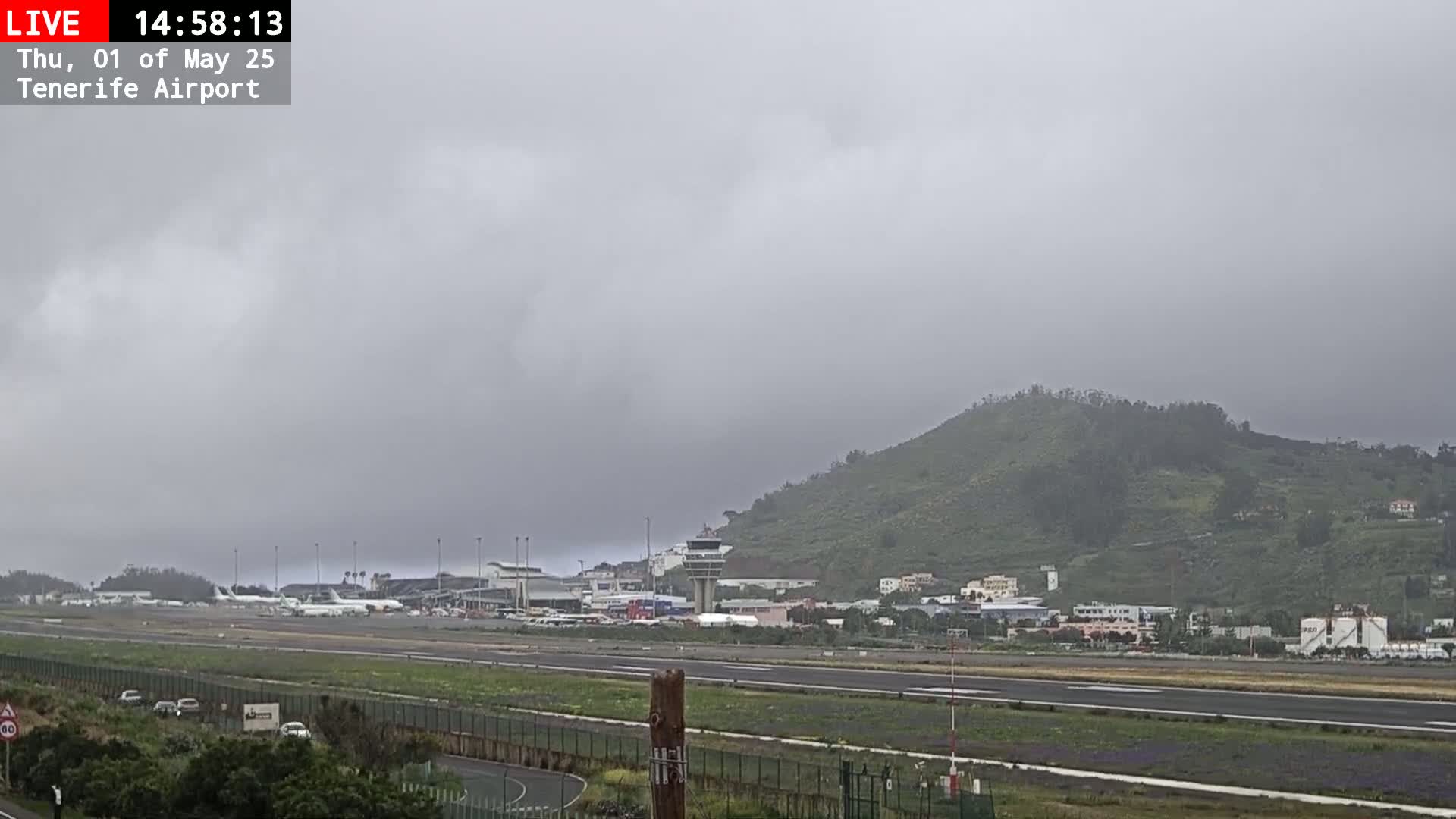 A mostly cloudy, overcast day at an airport with several airplanes parked on the tarmac, in front of a green hill.