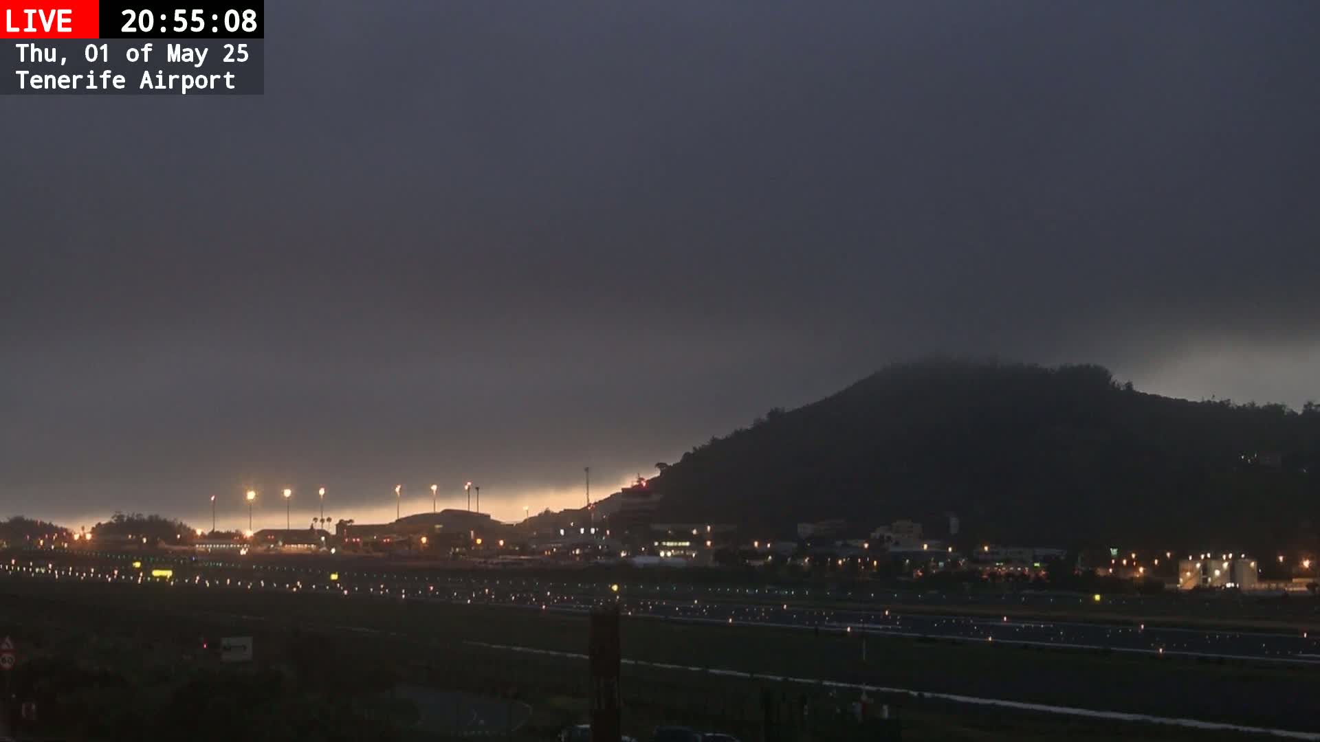 A dark, overcast night scene shows an airport runway with lights illuminating the tarmac, buildings, and a hill shrouded in fog or low clouds in the background.