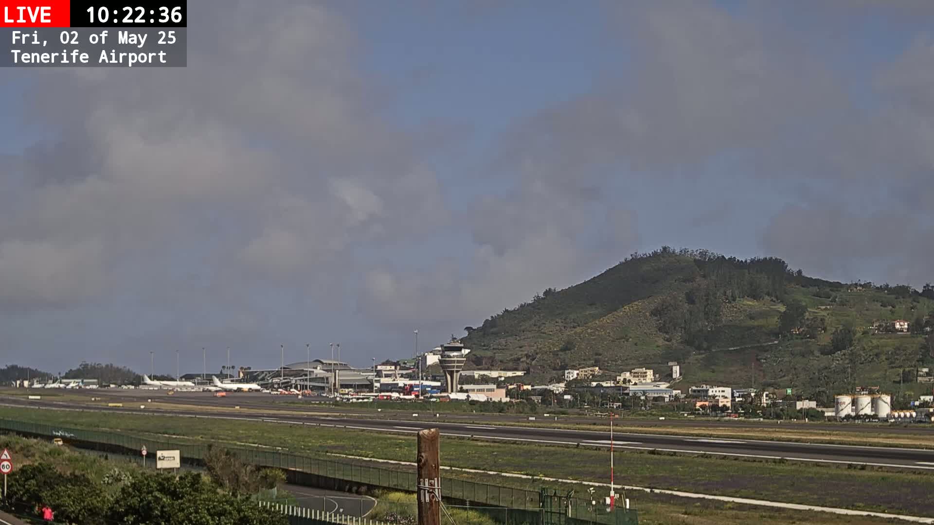An airport runway with several airplanes parked near the terminal buildings, under a partly cloudy sky, is situated at the foot of a green, hilly landscape.