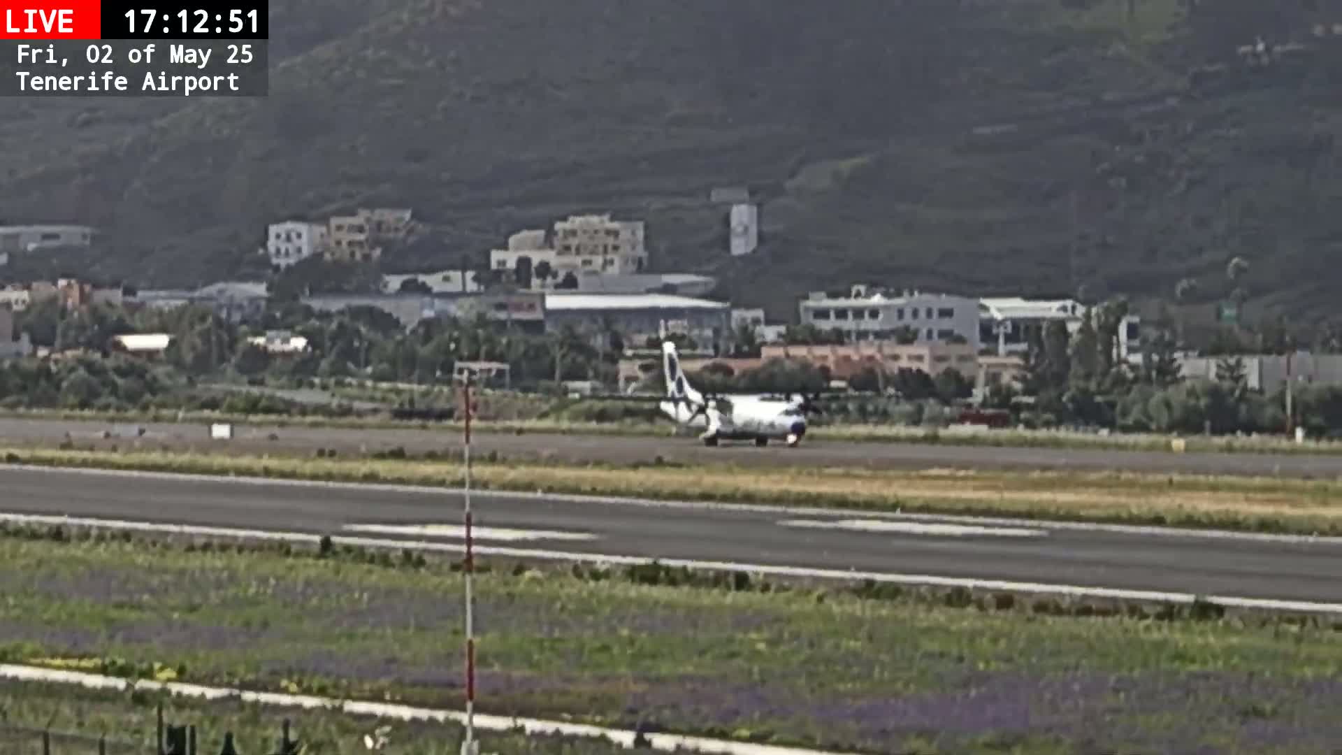 A small airplane taxis on an airport runway near a hillside town under overcast skies.