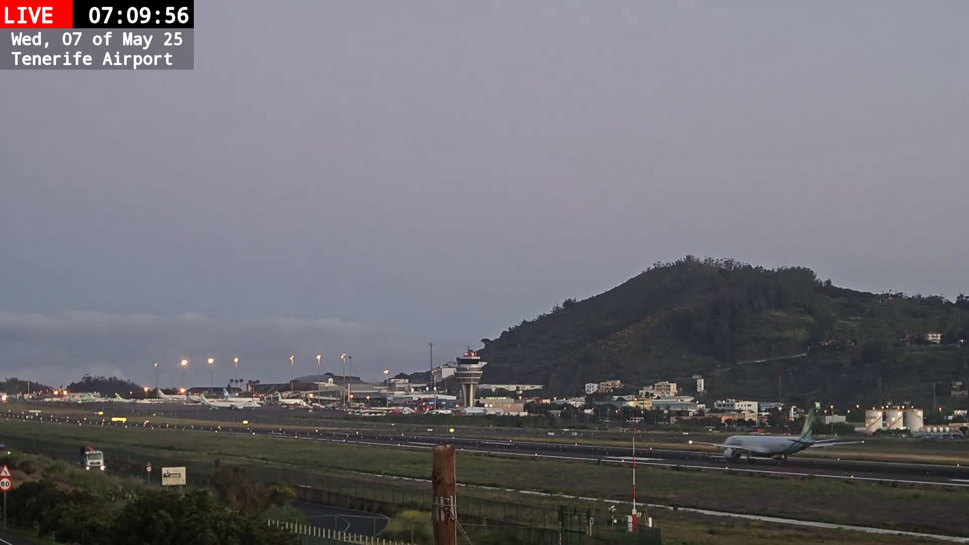 An airplane taxis on a runway at an airport near a hill under an overcast sky.