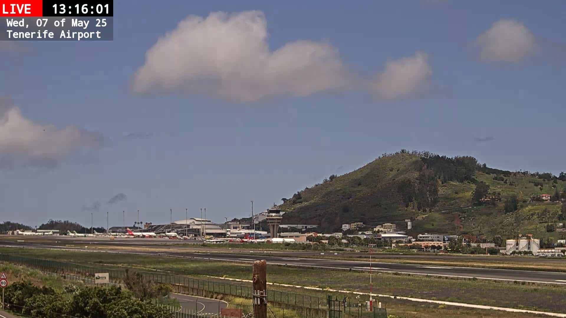 A sunny day with partly cloudy skies shows an airport runway with several airplanes, surrounded by low-lying green hills.