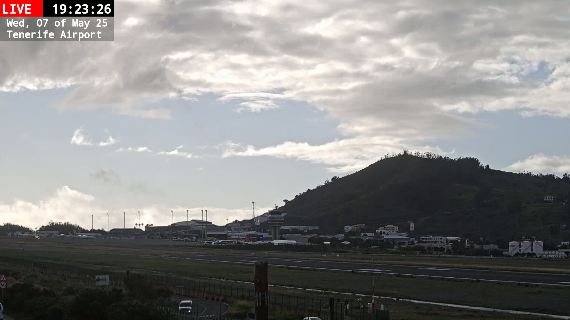 A mostly cloudy sky hangs over an airport runway with a dark, verdant hill in the background.