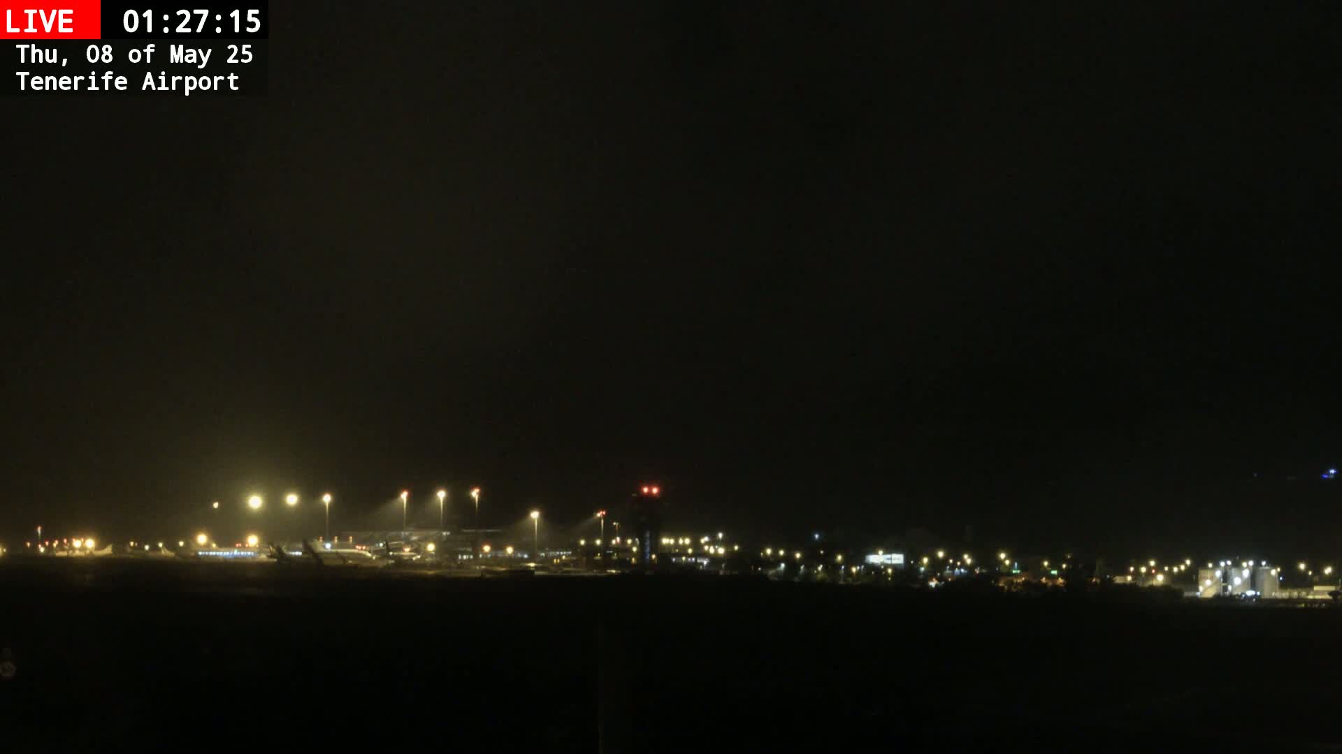A nighttime long shot of an airport runway and terminal buildings illuminated by lights under a dark, overcast sky.