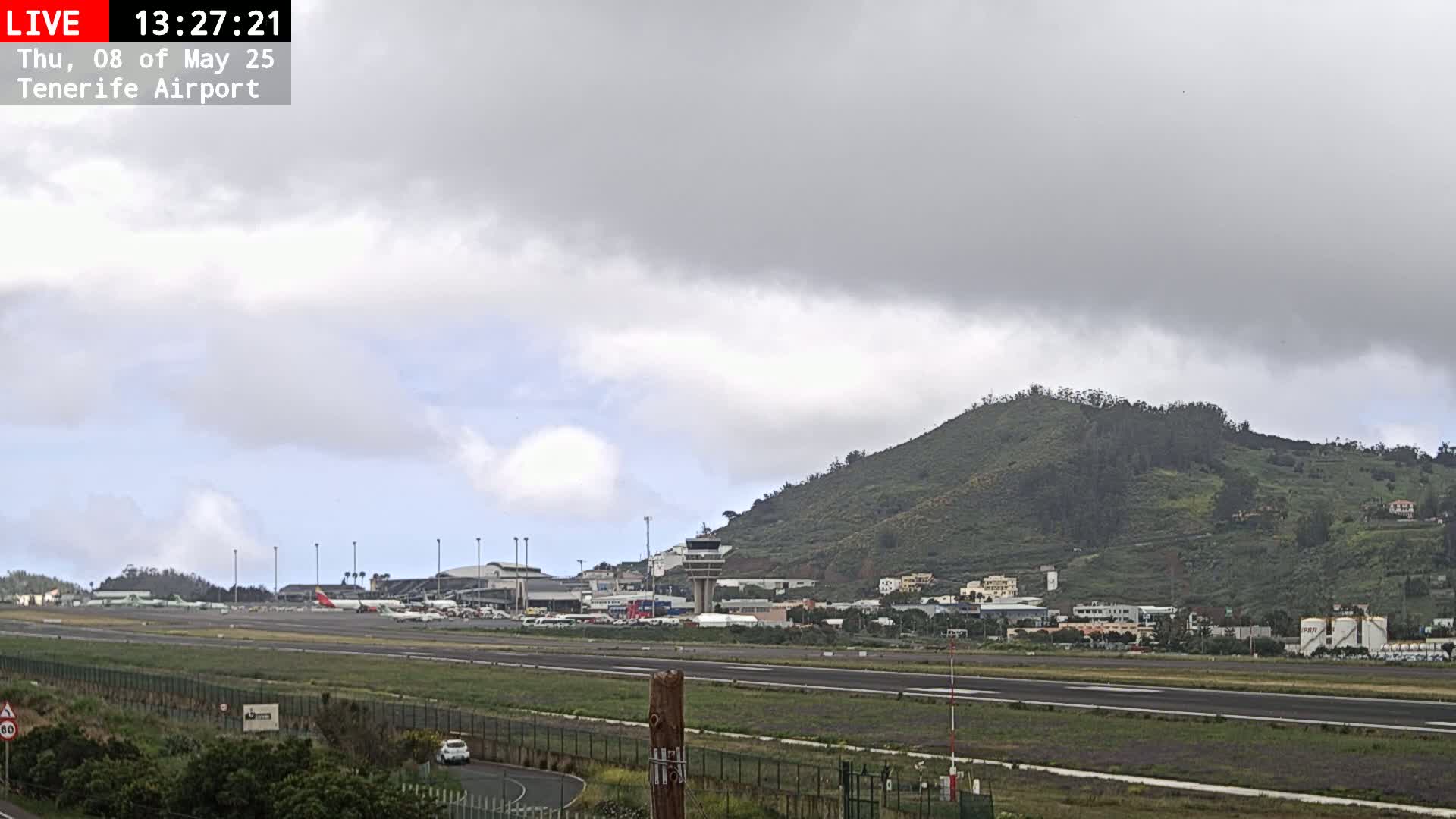 An airport runway with several planes parked on the tarmac, a control tower, and a green hill in the background under an overcast sky.