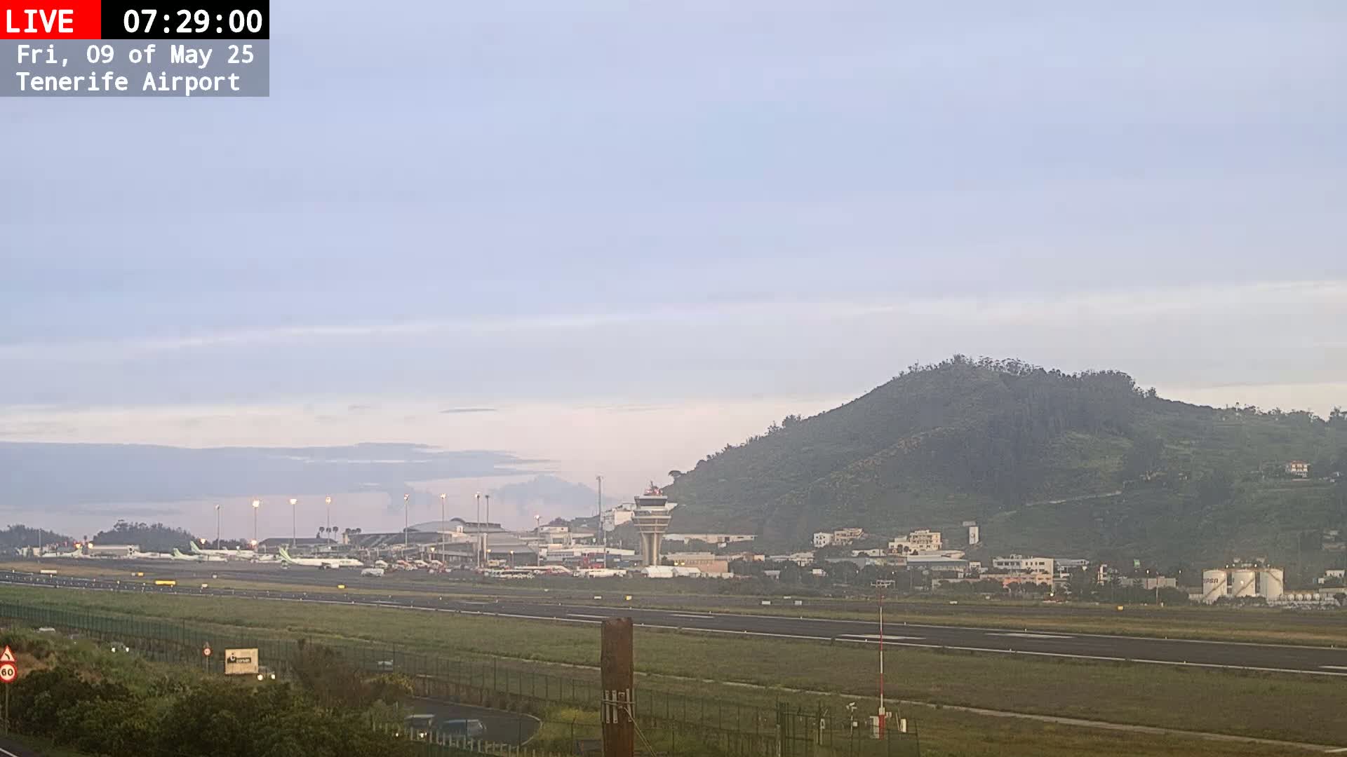 A long-range view of an airport runway, terminal buildings, and control tower under a hazy, light-blue sky, with a large hill in the background.