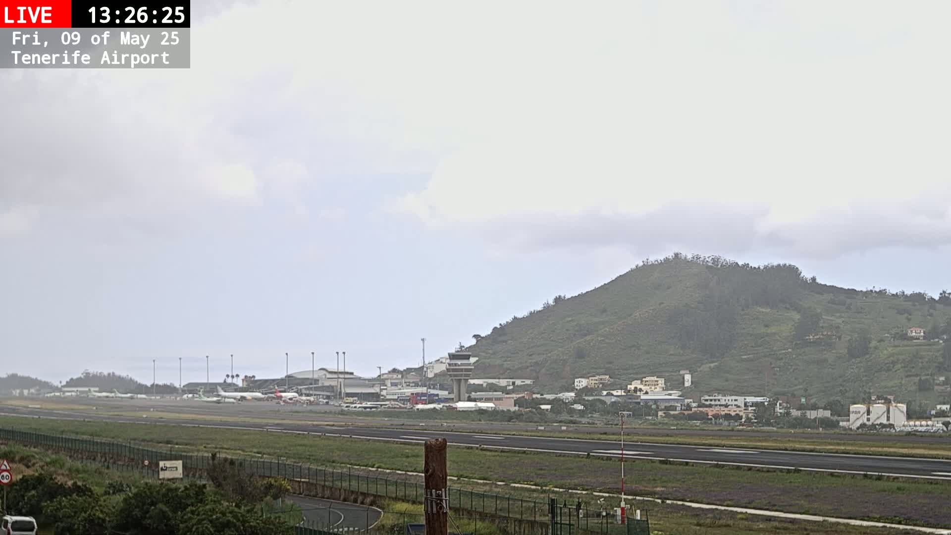 An airport runway and terminal are visible beneath an overcast sky, with a verdant hill rising in the background.