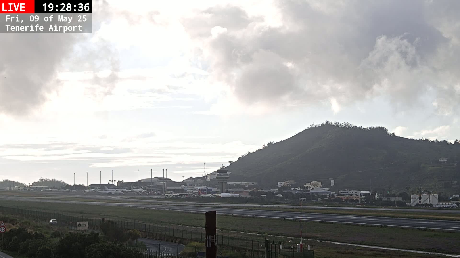 A mostly cloudy sky overlooks an airport runway with several planes parked, some buildings, and a large, dark hill in the background.