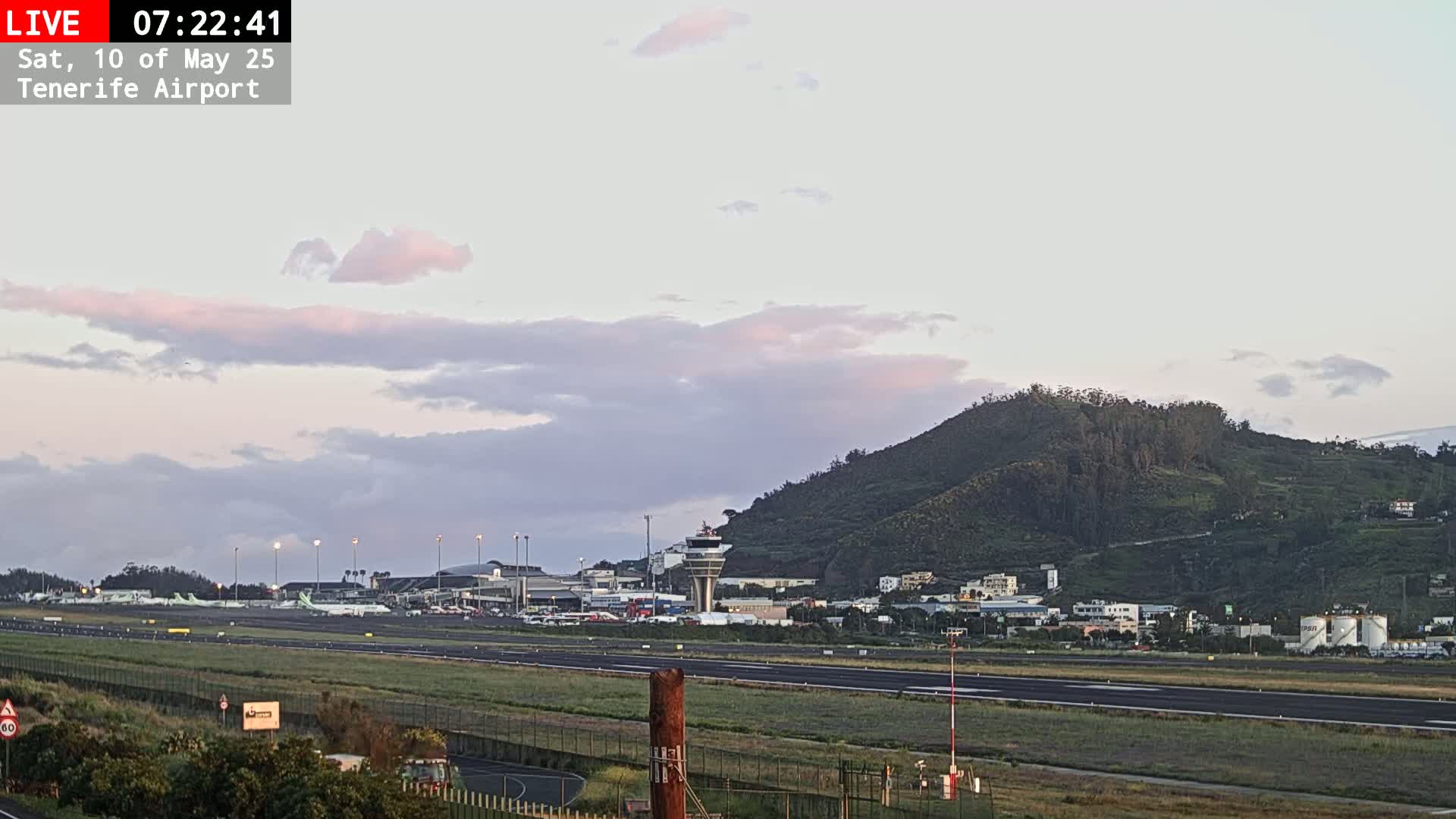 A view of an airport runway with several airplanes parked, a control tower, and a large green hill in the background under a partly cloudy, pale sky.