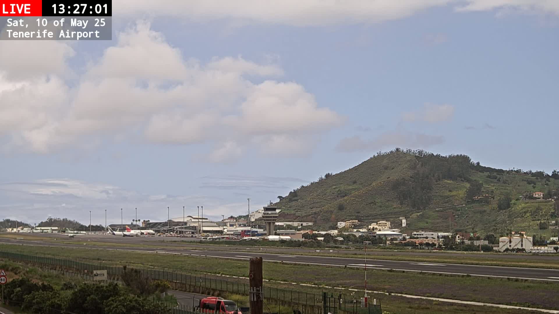A partly cloudy sky overlooks an airport runway with several planes, buildings, and a hill in the background.
