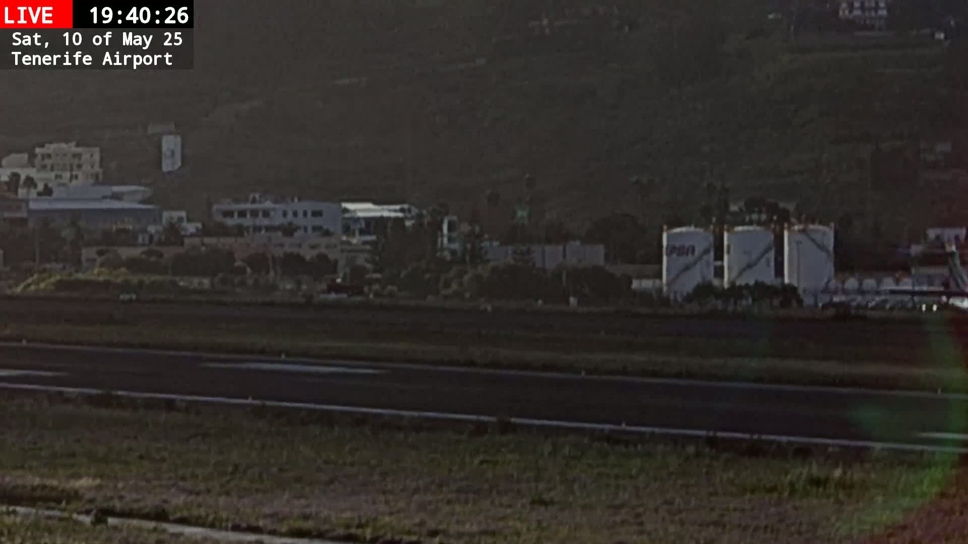A runway at an airport is visible, with buildings and fuel tanks in the background under dim, dusky lighting.