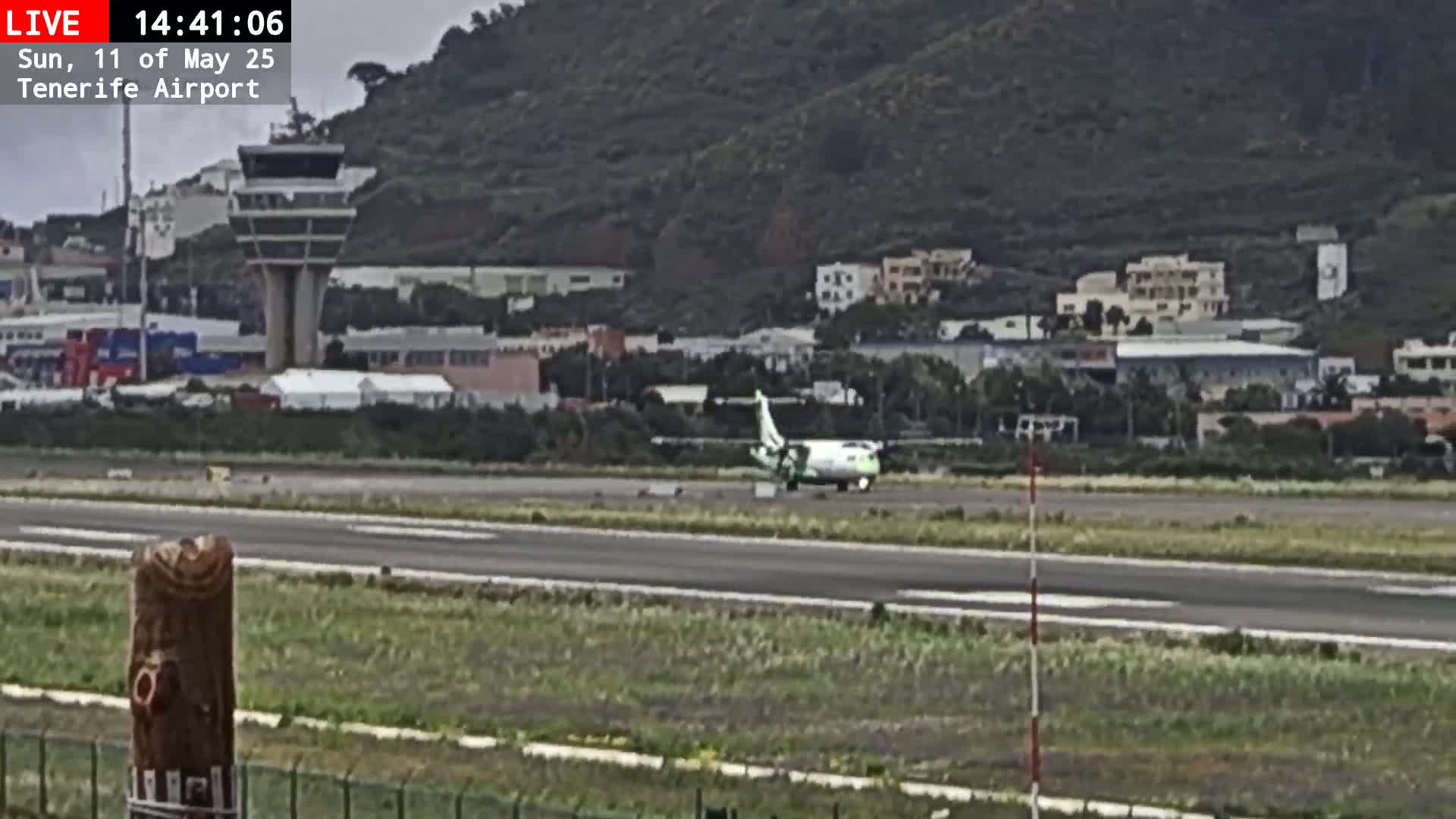 A small passenger plane is taxiing on an airport runway under overcast skies, with a control tower and buildings visible in the background.
