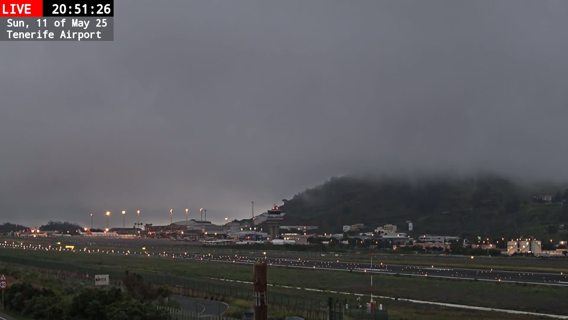 A foggy, dark nighttime scene shows an airport runway with buildings and hills in the background.