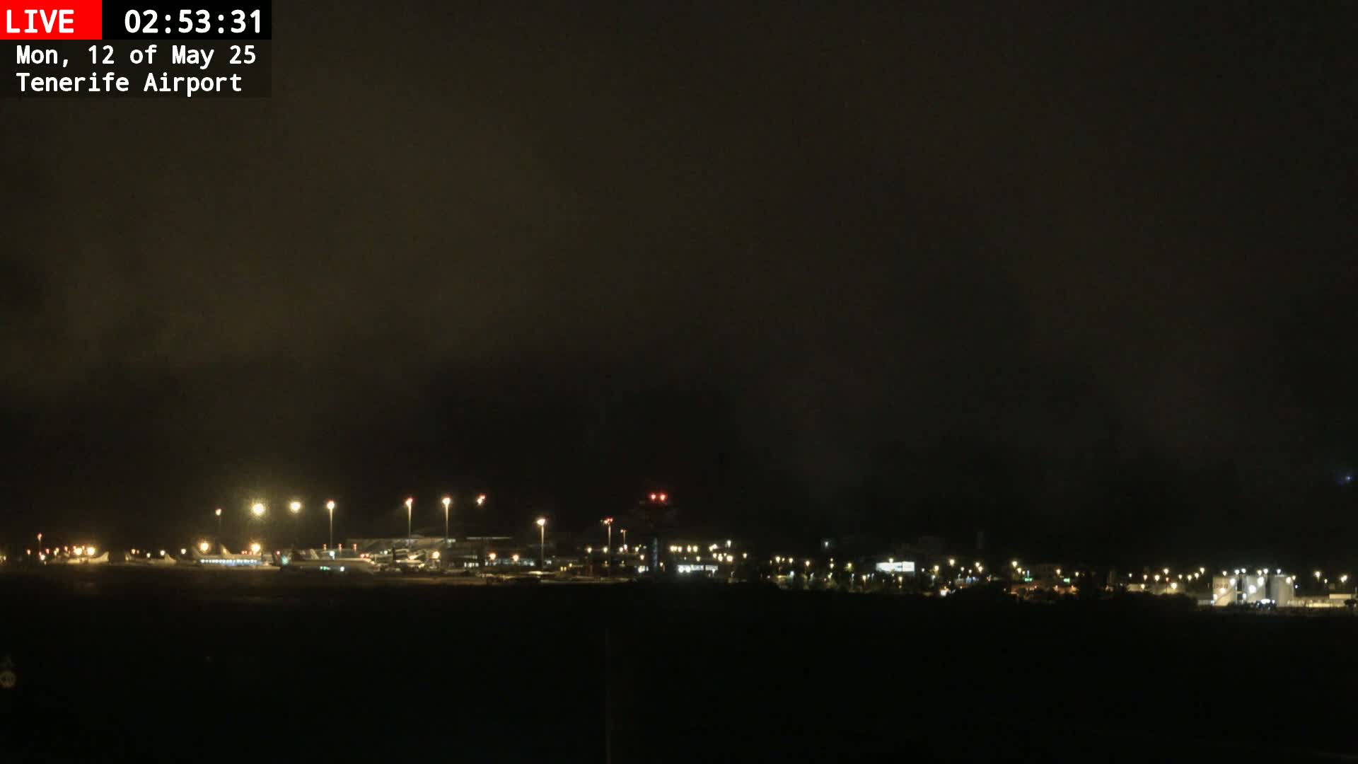 A nighttime view of an airport under a dark, cloudy sky, showing illuminated buildings and runways.