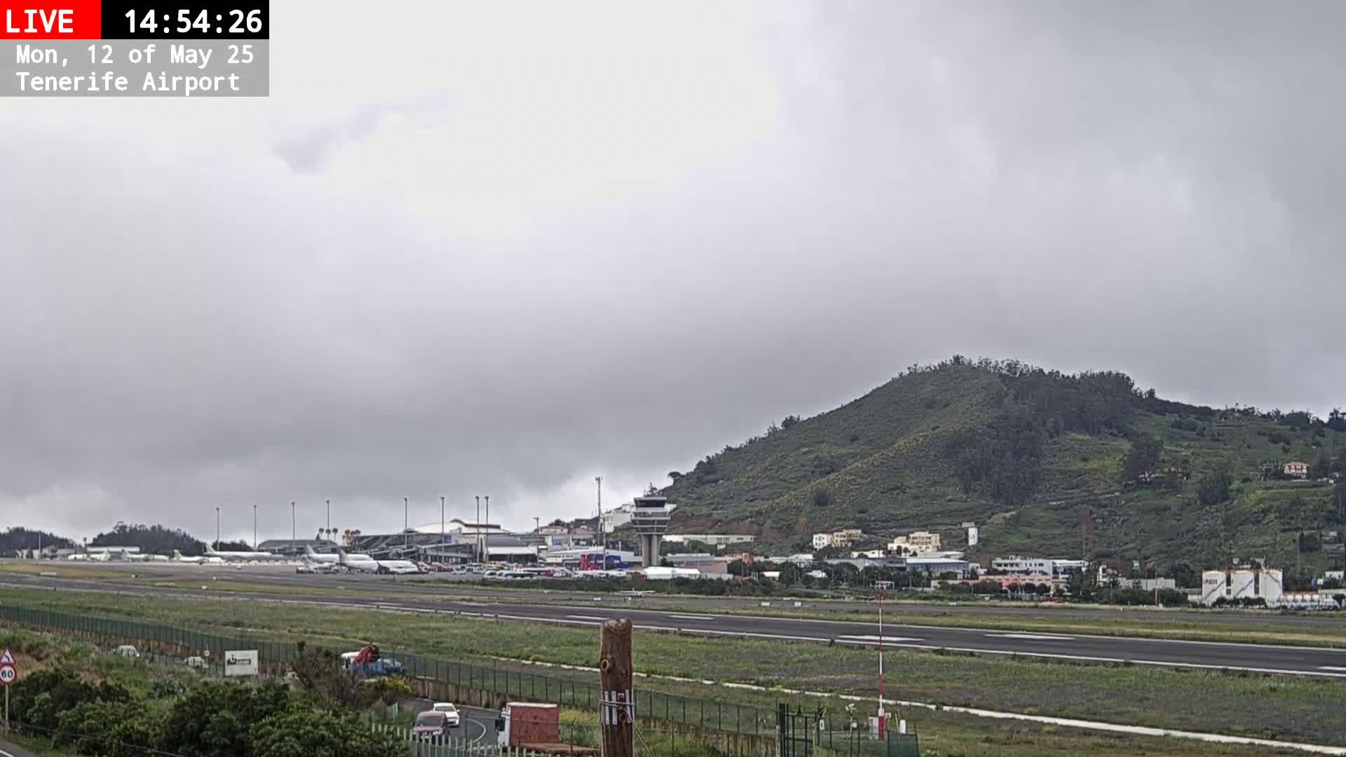 A cloudy day at an airport, showing multiple airplanes parked on the tarmac, airport buildings, and a green hill in the background.