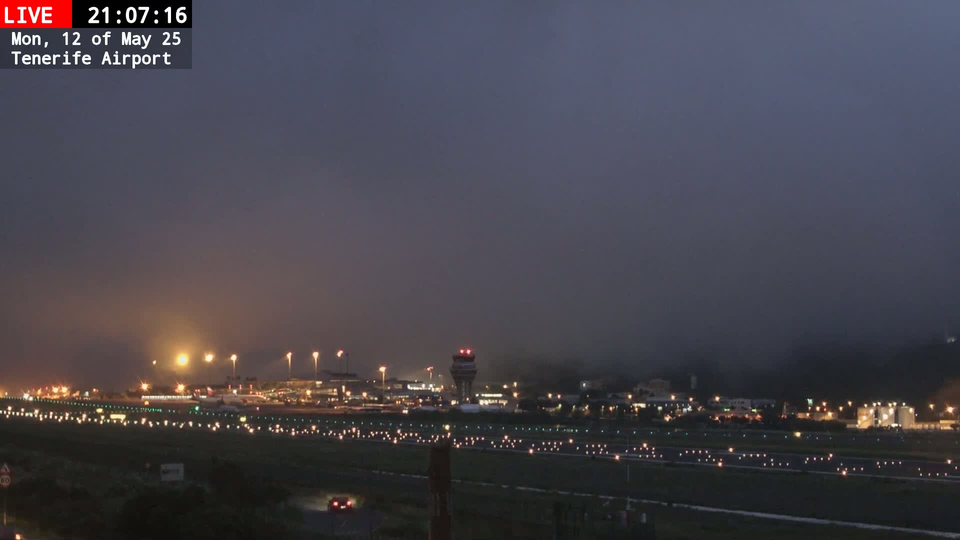 A foggy night scene shows an airport runway with illuminated lights and a control tower, surrounded by dimly lit buildings and vegetation.