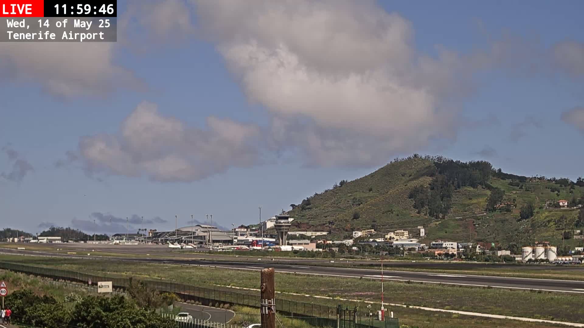 A partly cloudy day shows an airport runway, terminal buildings, and a green hill in the background.