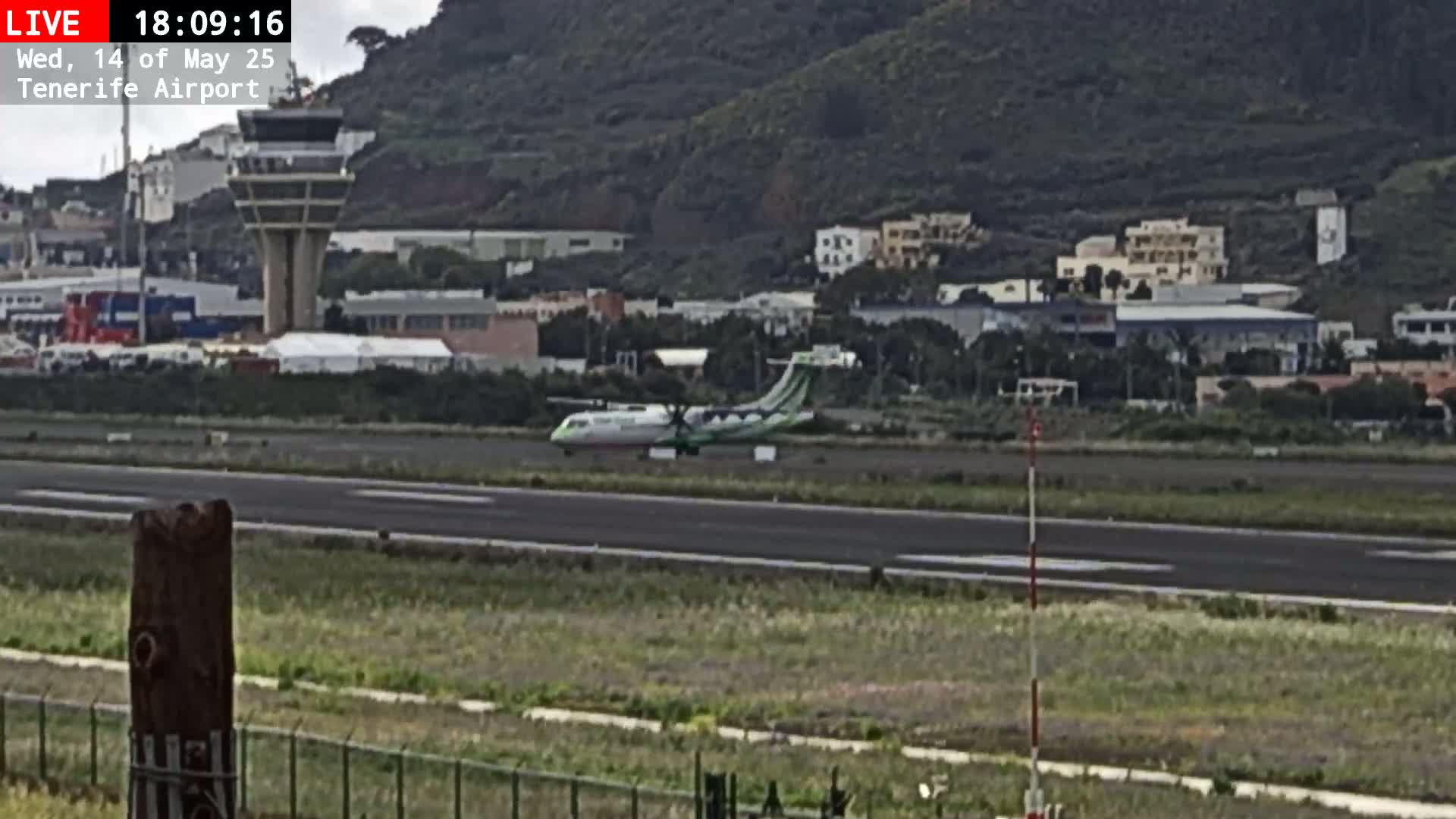 A turboprop airplane taxis on an airport runway next to a control tower, with a hilly, overcast background.