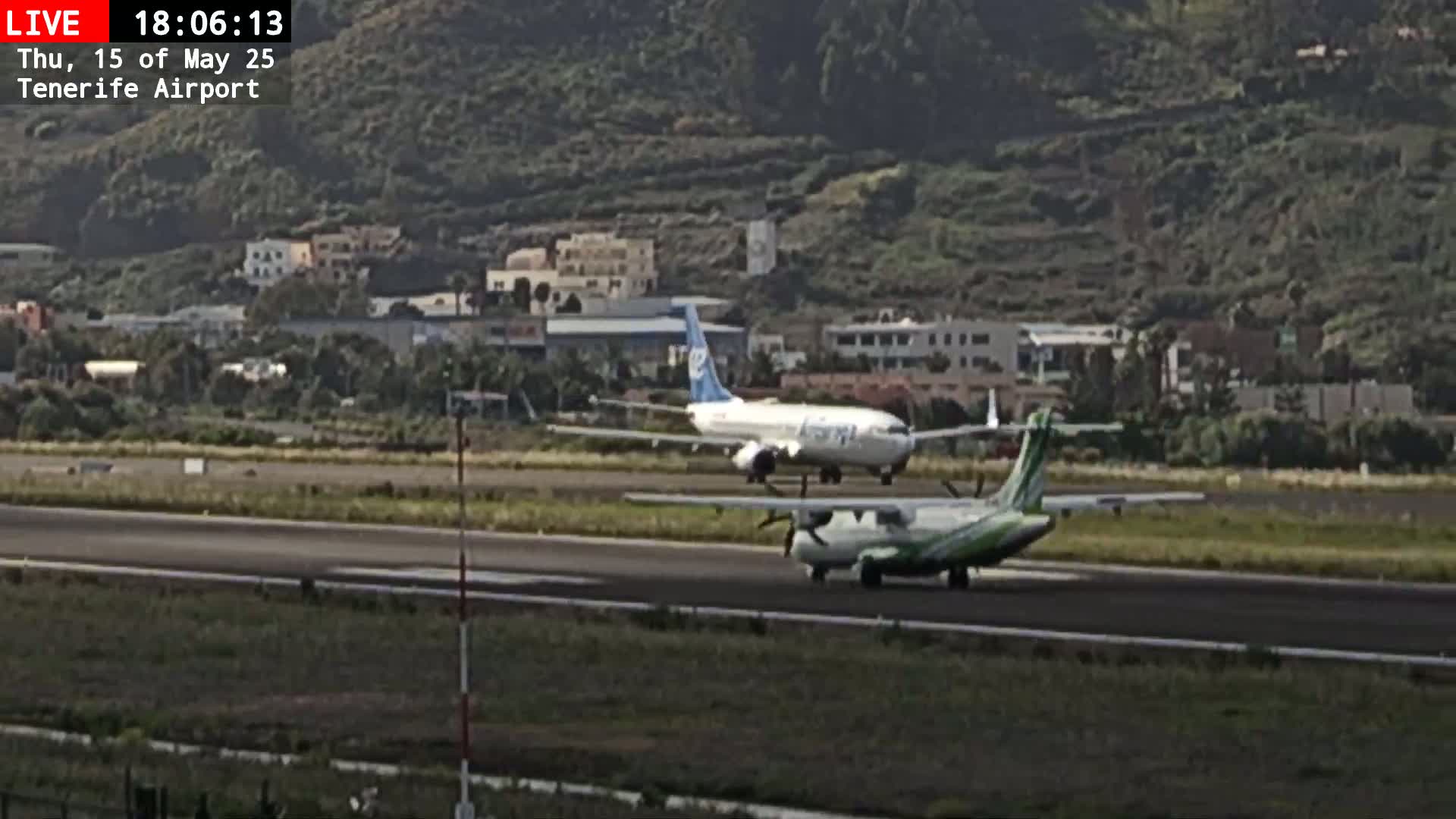 A smaller, green-and-white turboprop airplane taxis on an airport runway past a larger, blue-and-white jet airplane, with hills and buildings in the background under a mostly clear sky.