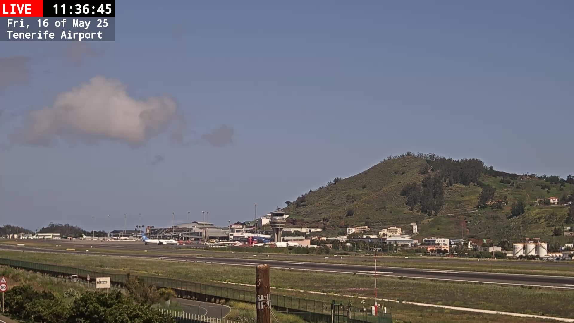 A mostly clear sky with a single puffy cloud hangs over an airport runway, with a plane visible on the tarmac and a green hill in the background.