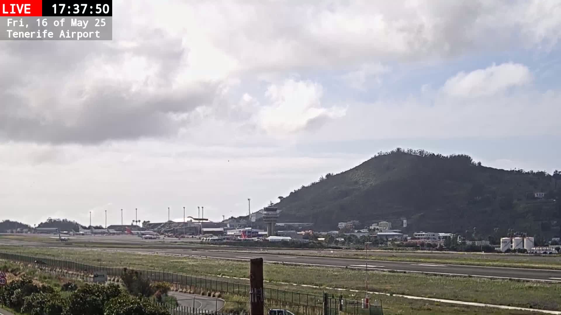 A mostly cloudy day at an airport, showing a runway, airplanes, airport buildings, and a large hill in the background.