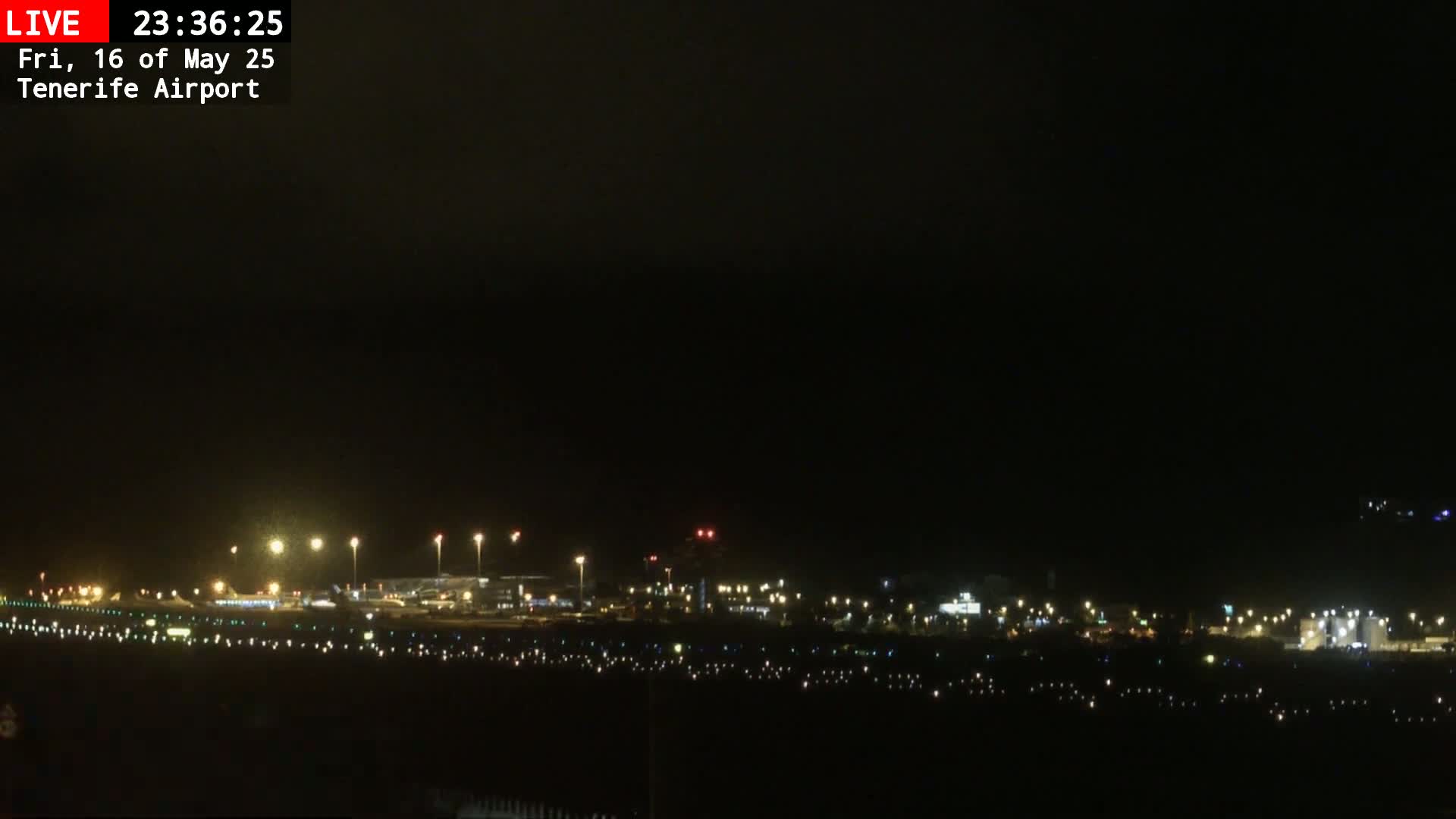 A nighttime long shot of an airport runway and terminal buildings, illuminated by numerous lights under a dark, cloudy sky.