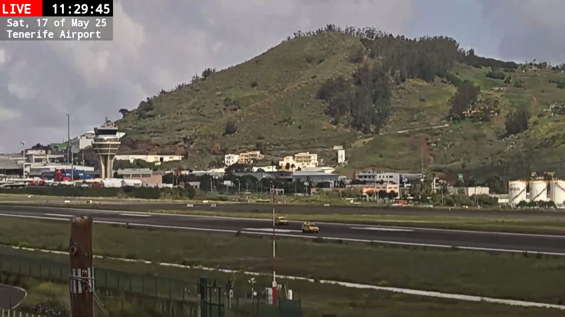 A partly cloudy day shows an airport runway with two yellow vehicles driving on it, near a control tower and a large green hill.