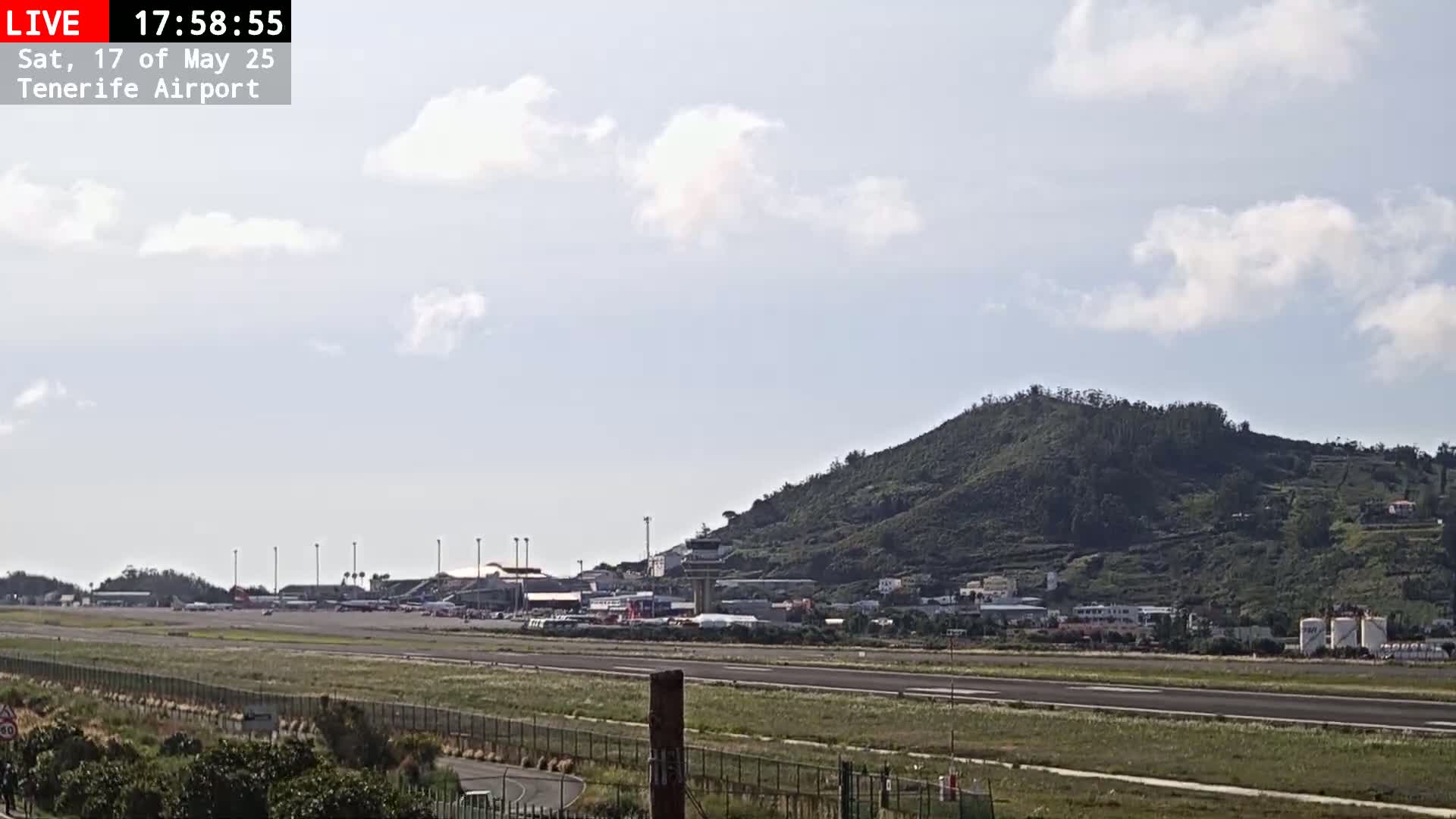 An airport runway stretches out before a verdant hill under a partly cloudy sky.
