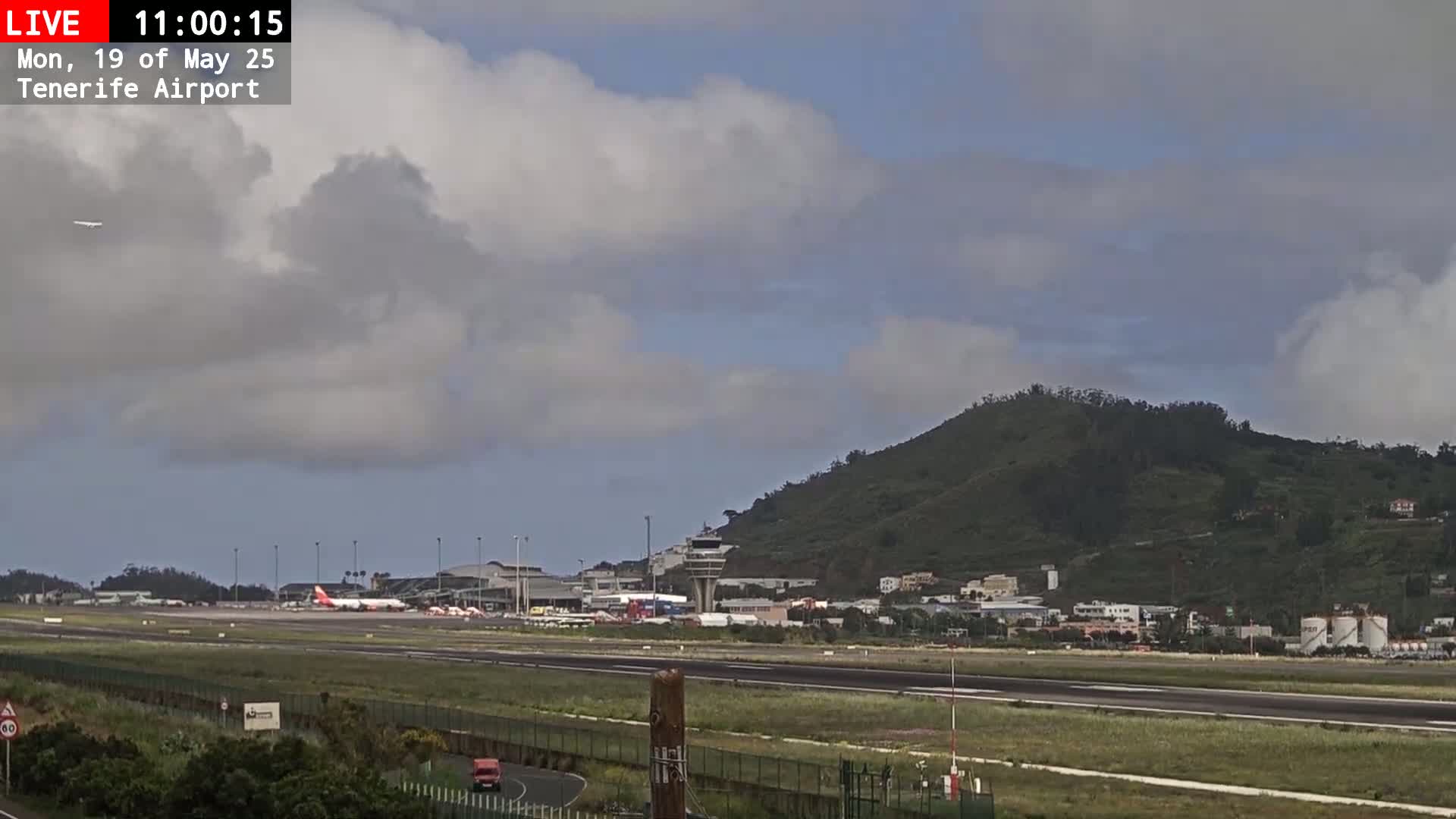 A mostly cloudy sky overlooks an airport runway with several planes and airport buildings, and a green hill in the background.