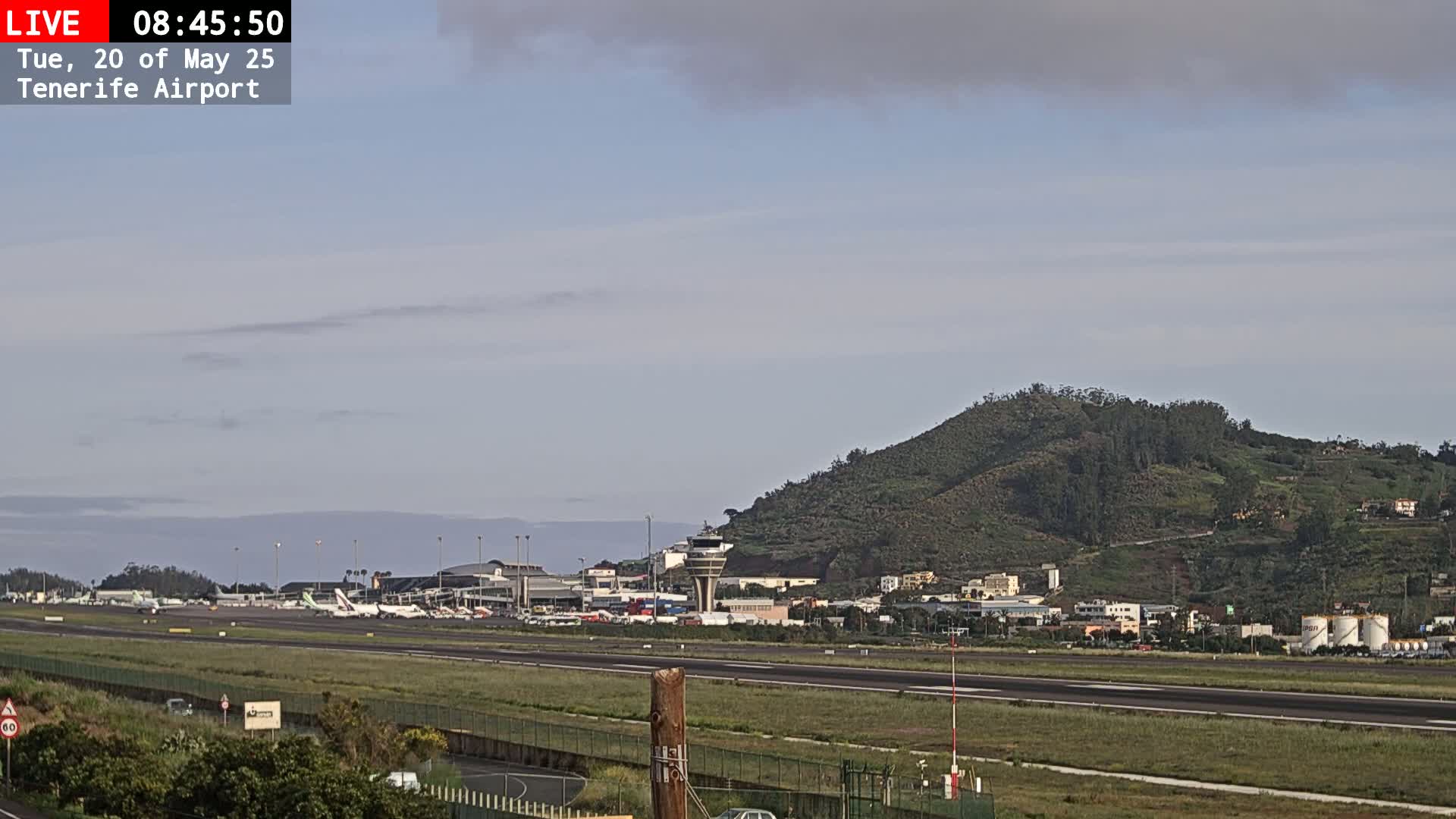 A partly cloudy day at Tenerife Airport shows airplanes parked on the tarmac, airport buildings, and a green hill in the background.