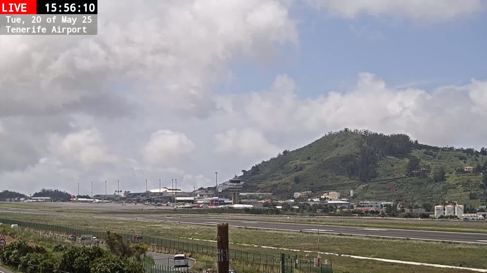 A partly cloudy day at an airport, showing runways, terminal buildings, and a green hill in the background.