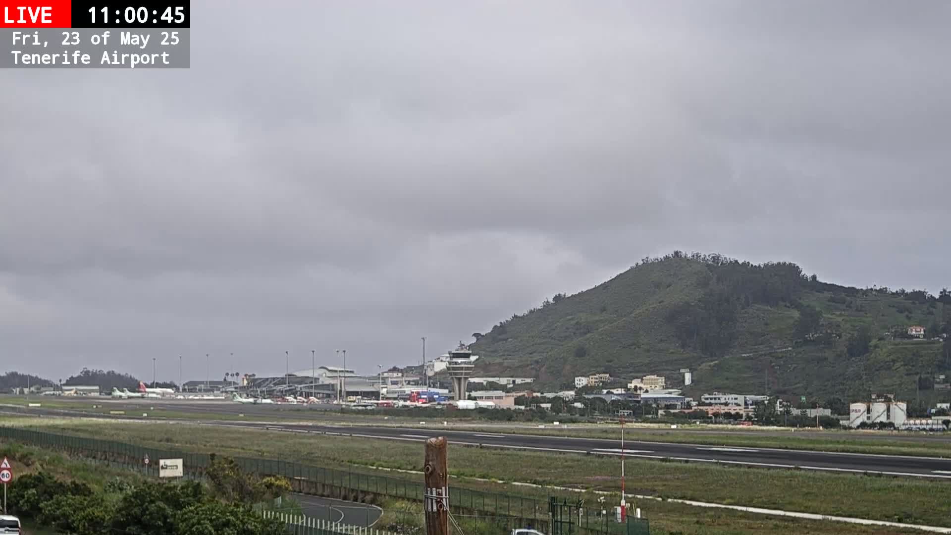 A view of an airport runway with several planes parked, adjacent buildings, and a verdant hill in the background under an overcast sky.