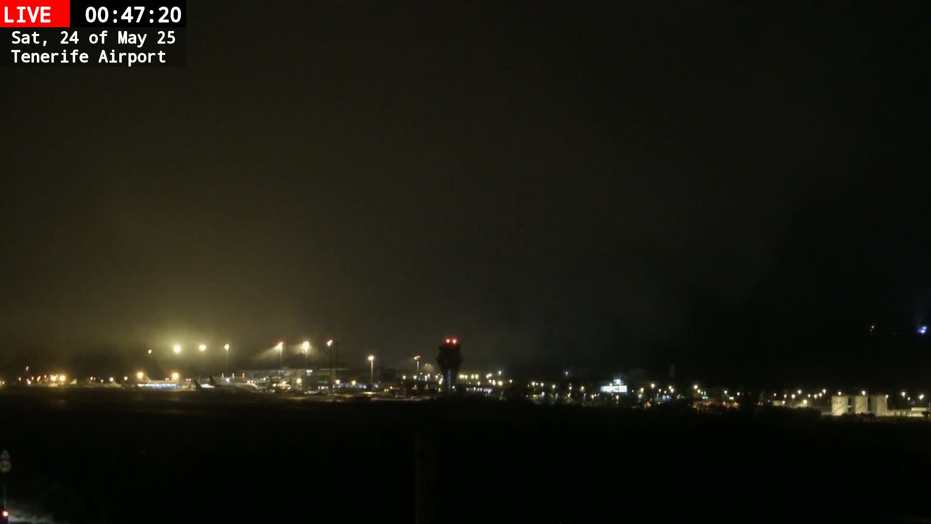 A nighttime view of an airport, dimly lit under hazy or foggy conditions, shows the control tower and runway lights.