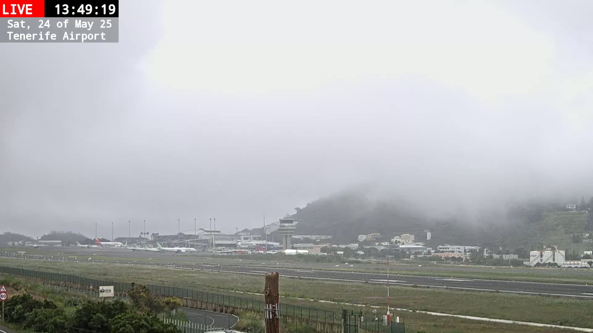 A foggy view of an airport runway with several airplanes parked near the terminal buildings and a control tower visible in the distance.