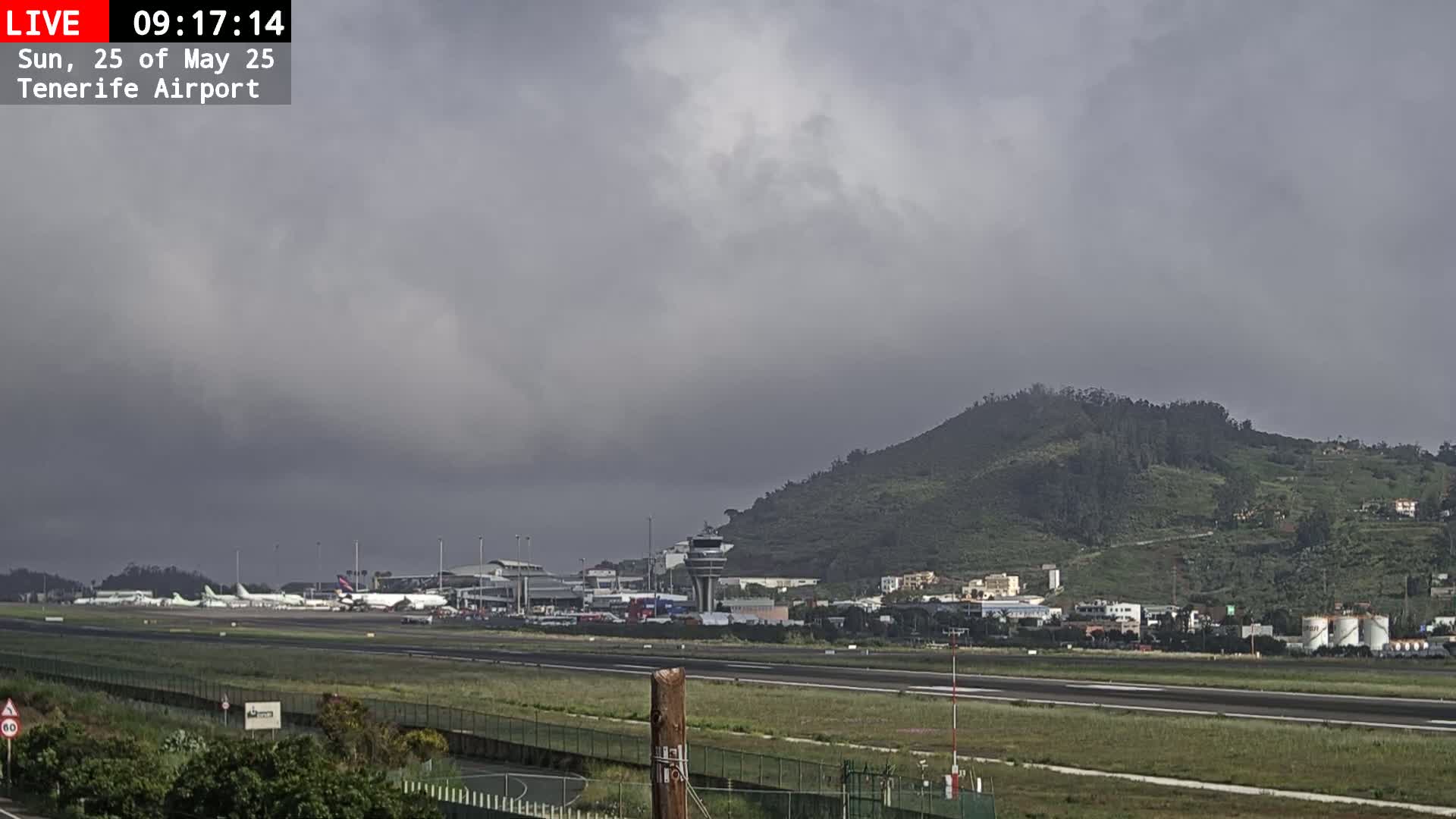 An overcast sky hangs over an airport with several airplanes parked on the tarmac, a control tower, and a verdant hill in the background.
