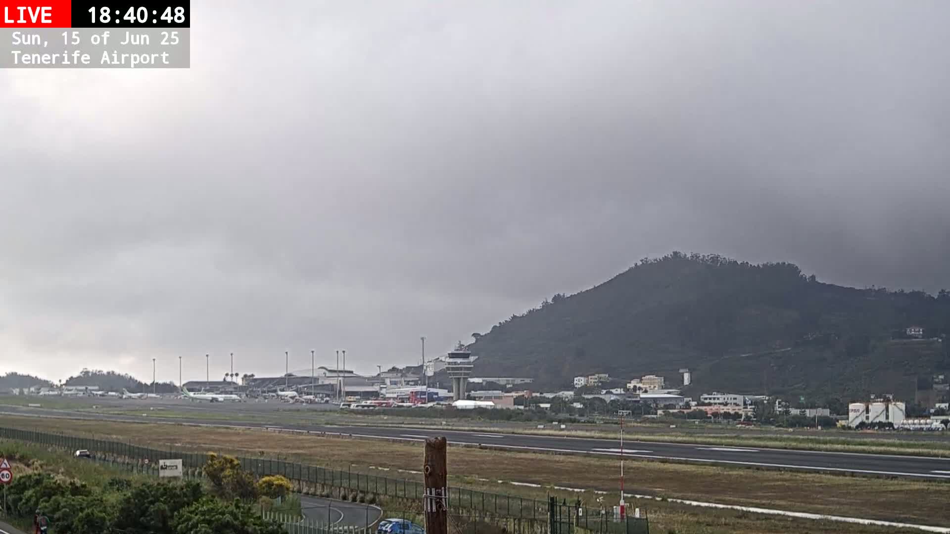 An airport runway and terminal are visible beneath an overcast sky, with a hill in the background.