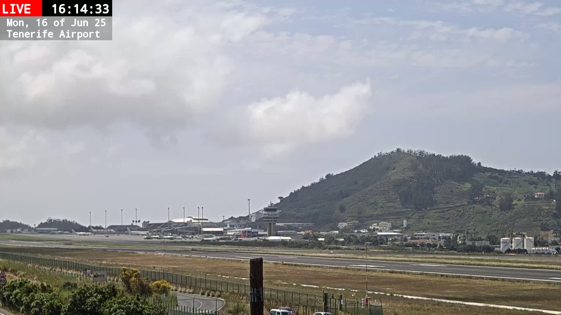 An airport runway, terminal buildings, and control tower are visible beneath a partly cloudy sky, with a large, green hill in the background.