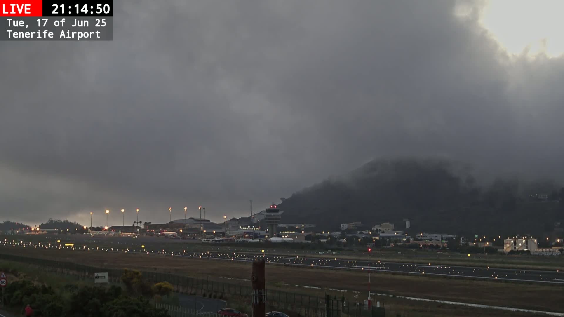 A mostly cloudy, overcast sky hangs over an airport runway with illuminated lights and airport buildings, and a hill shrouded in mist in the background.